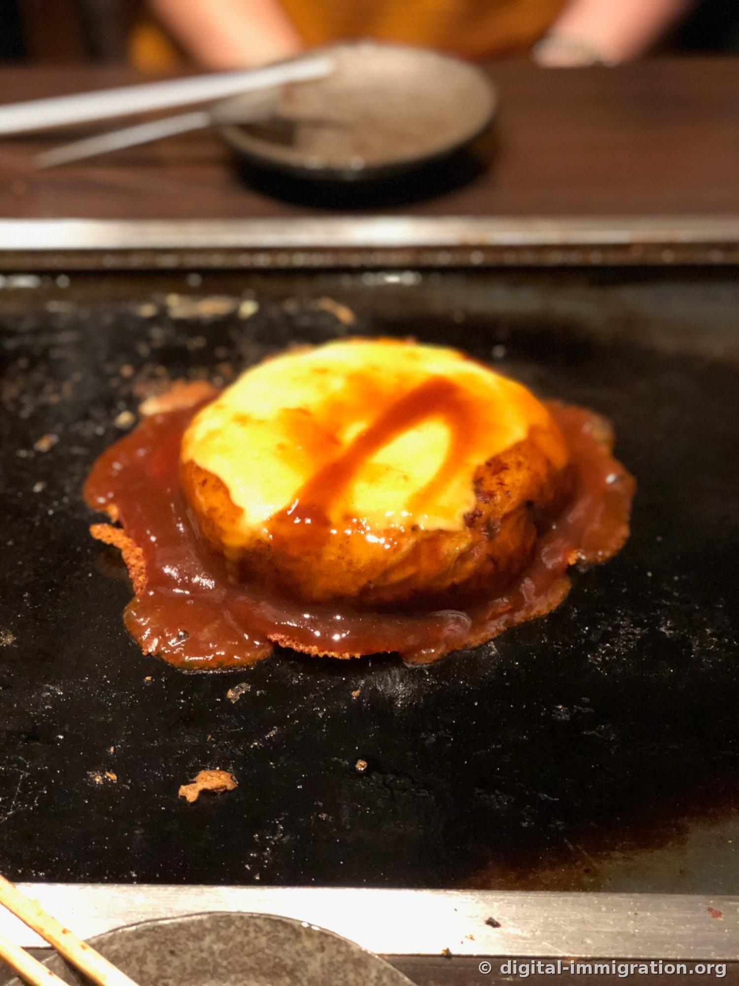 치즈를 덮은 오코노미야키 위에 갈색 소스를 지그재그로 뿌린 모습, 소스가 철판 위로 넓게 퍼져 있다