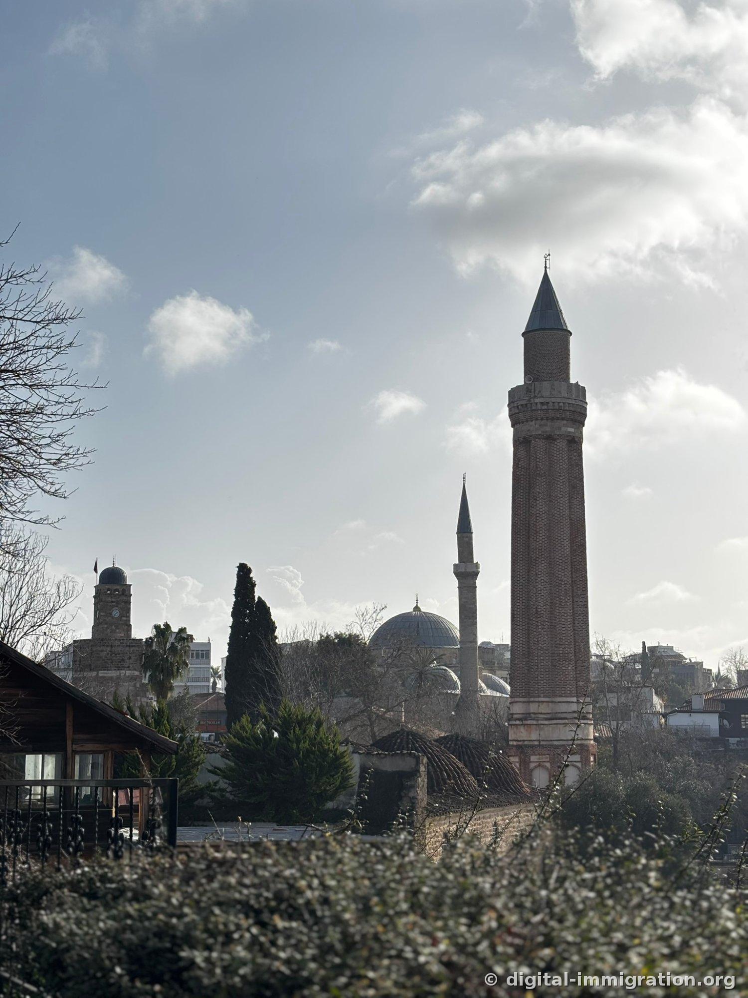 Under a cloudy sky, the red brick Yivli Minaret, a gray dome and white minaret to its left, and a clock tower in the far left distance together form the old city skyline.