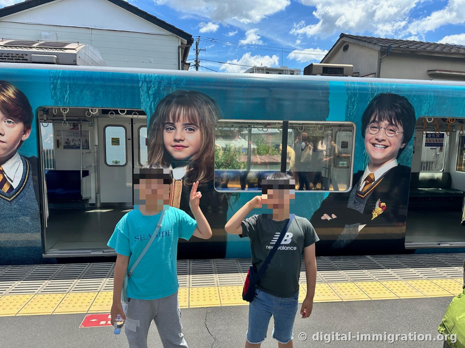 Two children making a V-sign in front of a blue train wrapped with Harry Potter characters on a platform