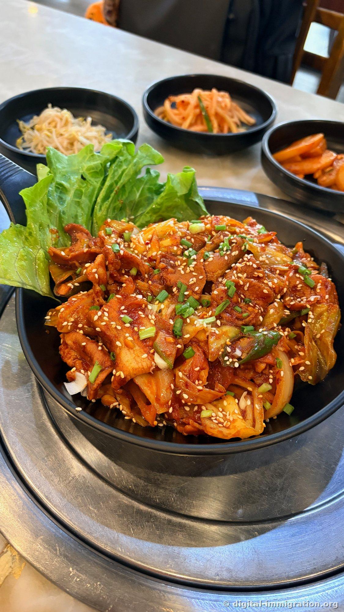 Jeyuk-bokkeum rouge dans un bol noir, avec oignon et chou sautés, parsemé de sésame et de ciboule, accompagné de laitue sur un côté et, en arrière-plan, de pousses de soja assaisonnées, de radis râpé assaisonné et de kimchi.