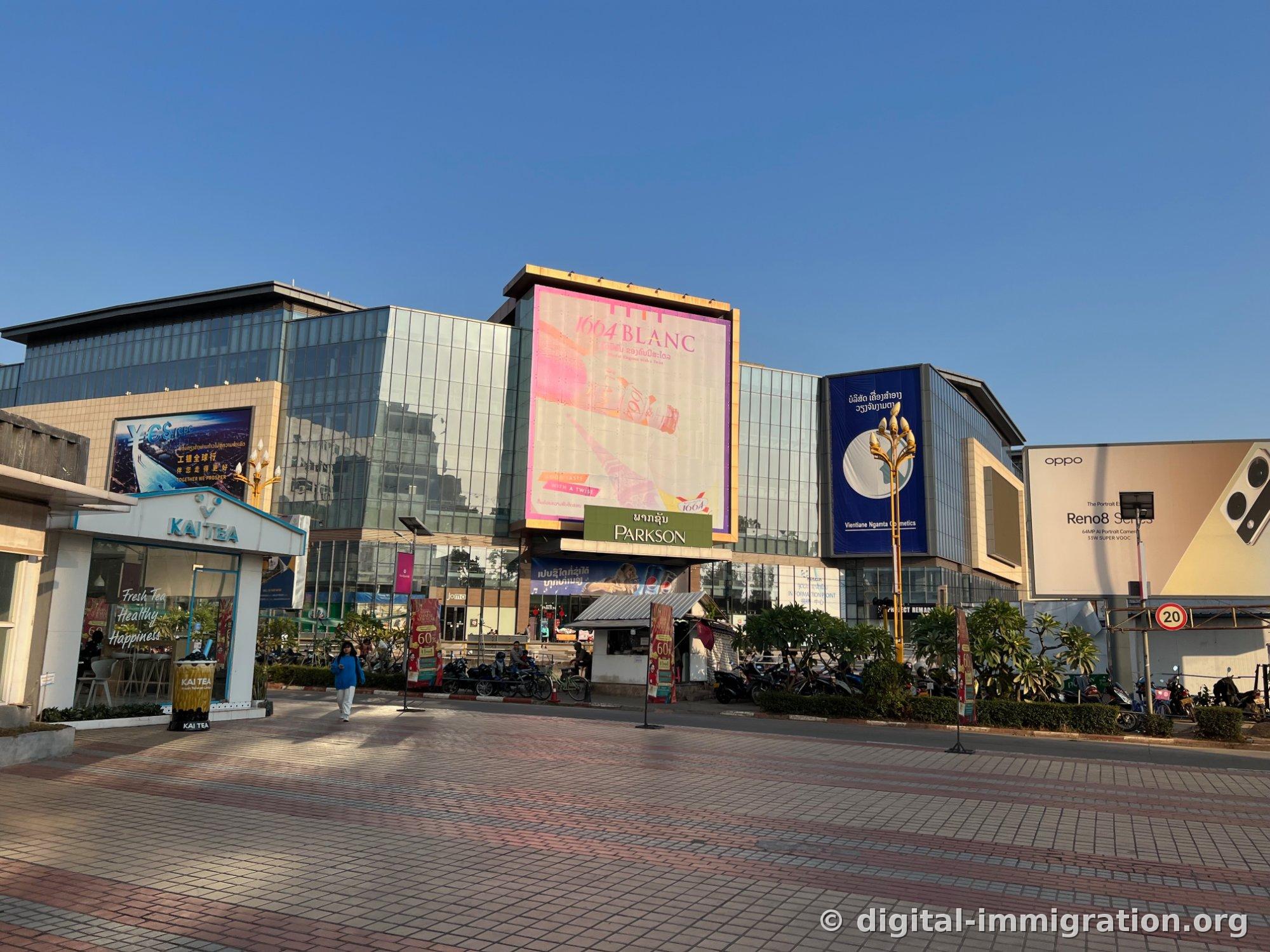 Vue d'ensemble du Parkson Mall de l'autre côté de la rue, avec le petit magasin KAI TEA à gauche et la publicité OPPO Reno8 à droite, avec des motos garées en ligne devant