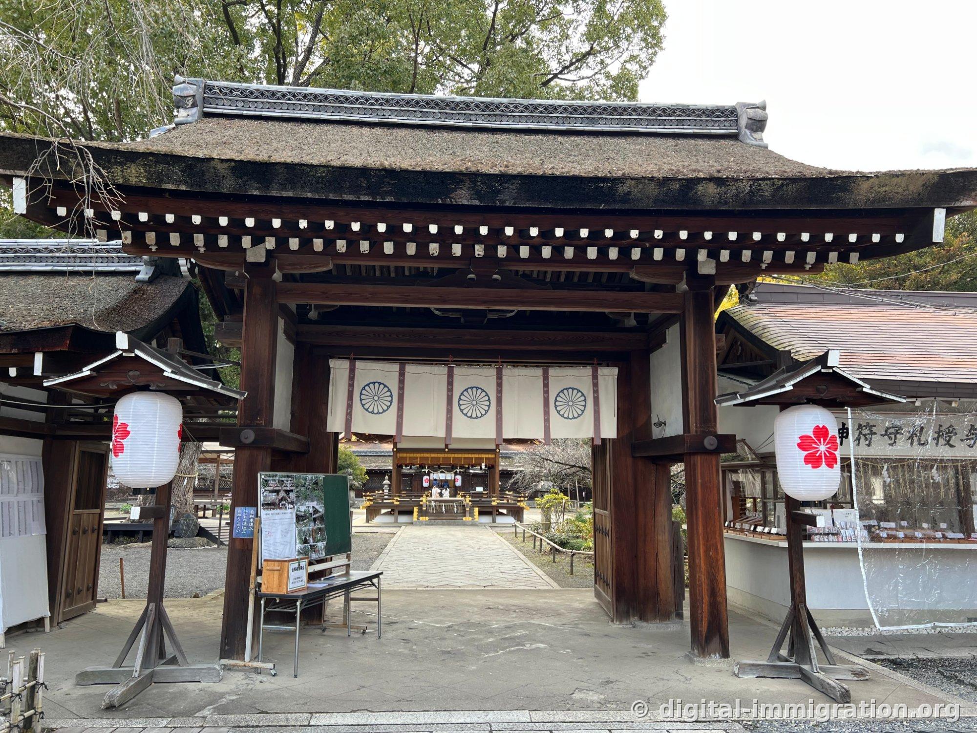 The south gate of Hirano Shrine with a moss-covered roof and dark wooden pillars, white curtain bearing the imperial chrysanthemum crest hanging from it, with white lanterns decorated with red cherry blossoms on both sides