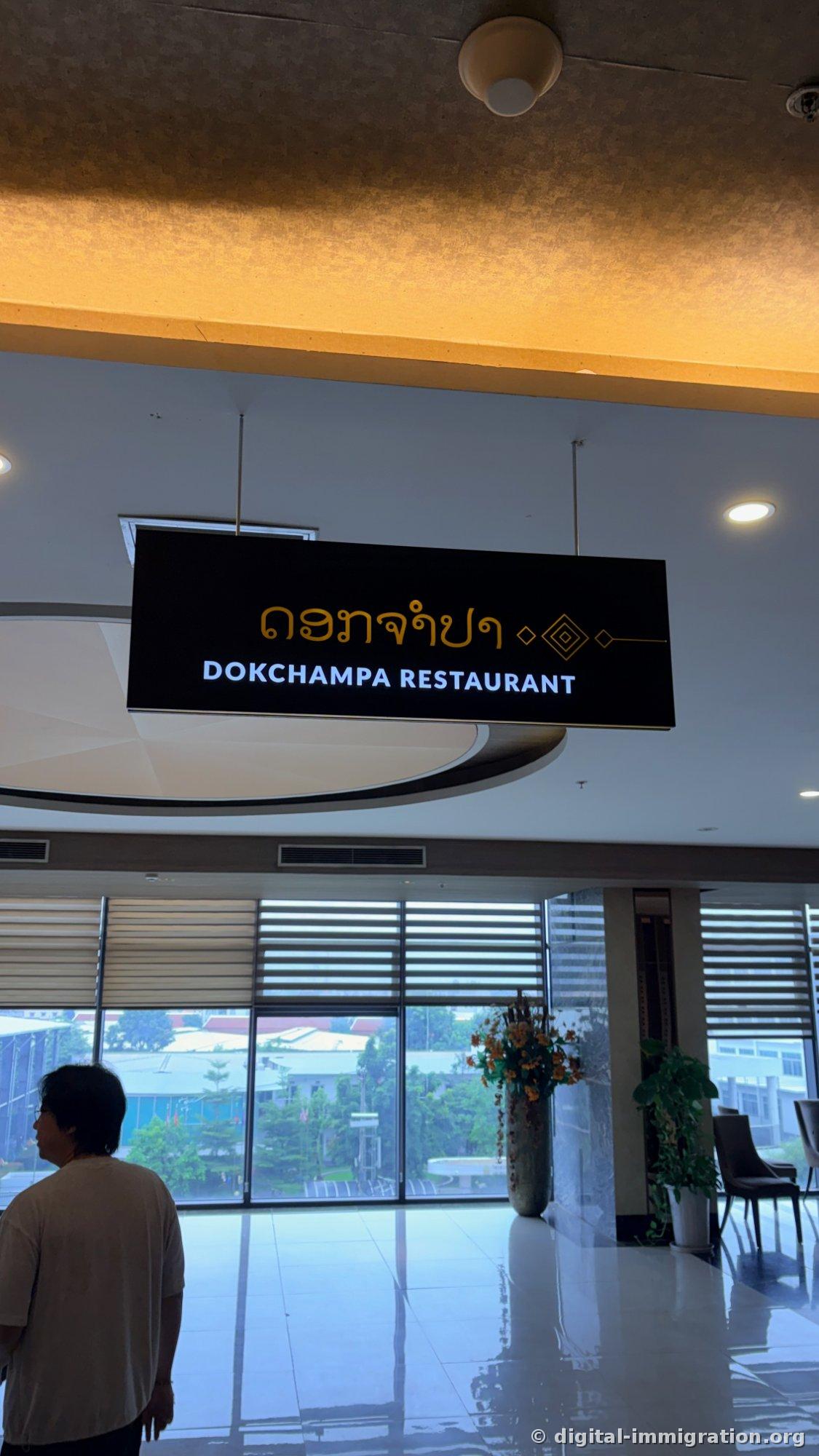 Enseigne du restaurant Dokchampa suspendue au plafond du lobby avec du texte en or en laotien et en blanc en anglais « DOKCHAMPA RESTAURANT », et la vue sur le jardin à travers la baie vitrée