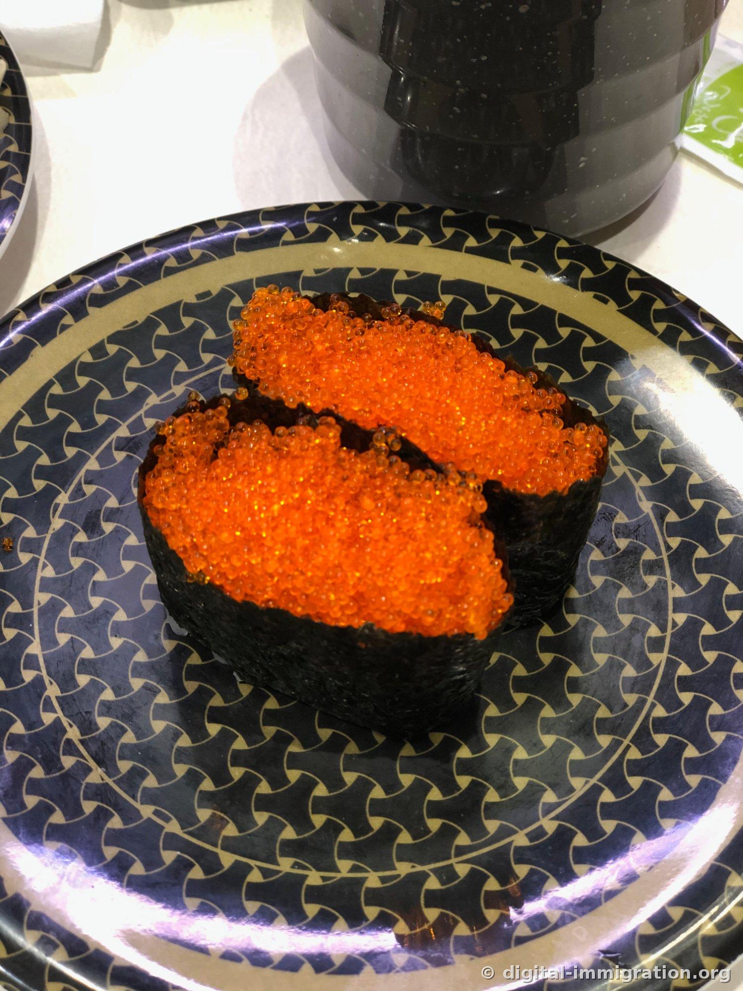 Sur une assiette avec un motif géométrique bleu marine, deux temaki d'œufs de poisson volant encerclés d'algue sont disposés légèrement avec une couleur orange