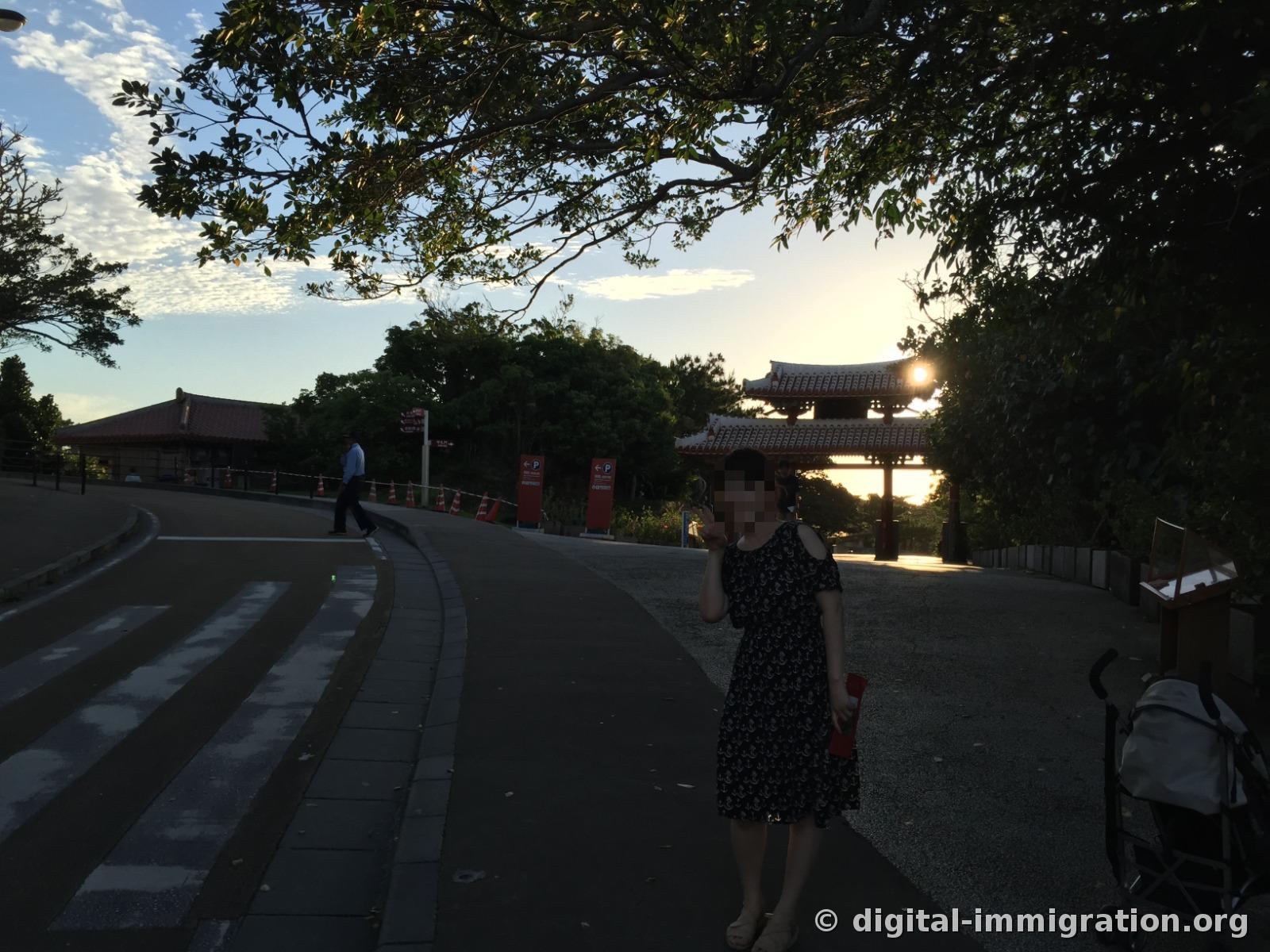 Shuri Castle park entrance road at sunset, the sun hanging above the wooden gate tower roof, parking sign and stroller on the roadside