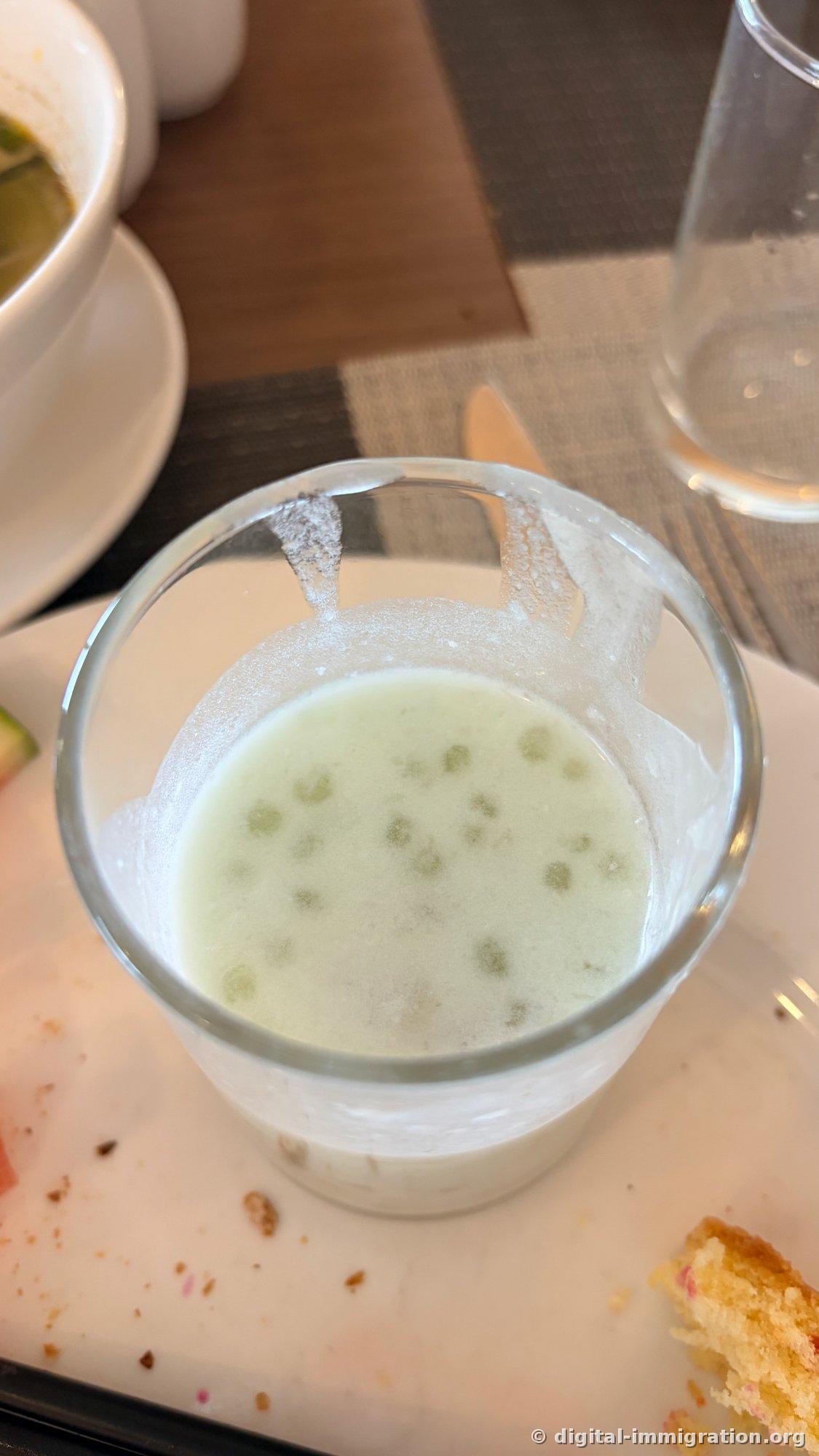 Verre de jus de légumes vert clair sur la table du petit-déjeuner, avec une mousse blanche collée à l'intérieur du verre, une assiette blanche à côté et des miettes de pain et un verre d'eau