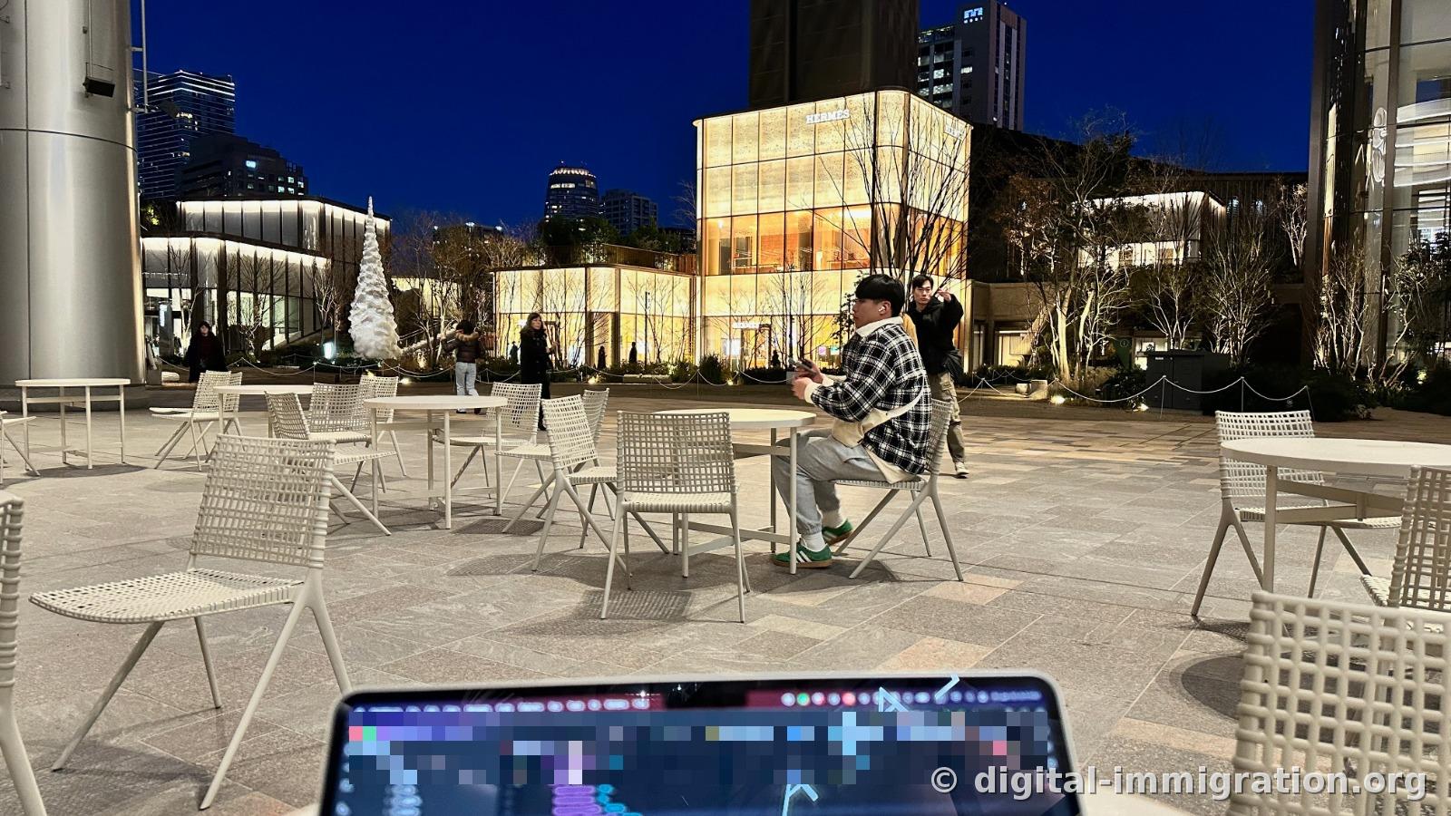 White tables and chairs in outdoor public seating, laptop screen in foreground, illuminated Hermès building and white conical sculpture across the way