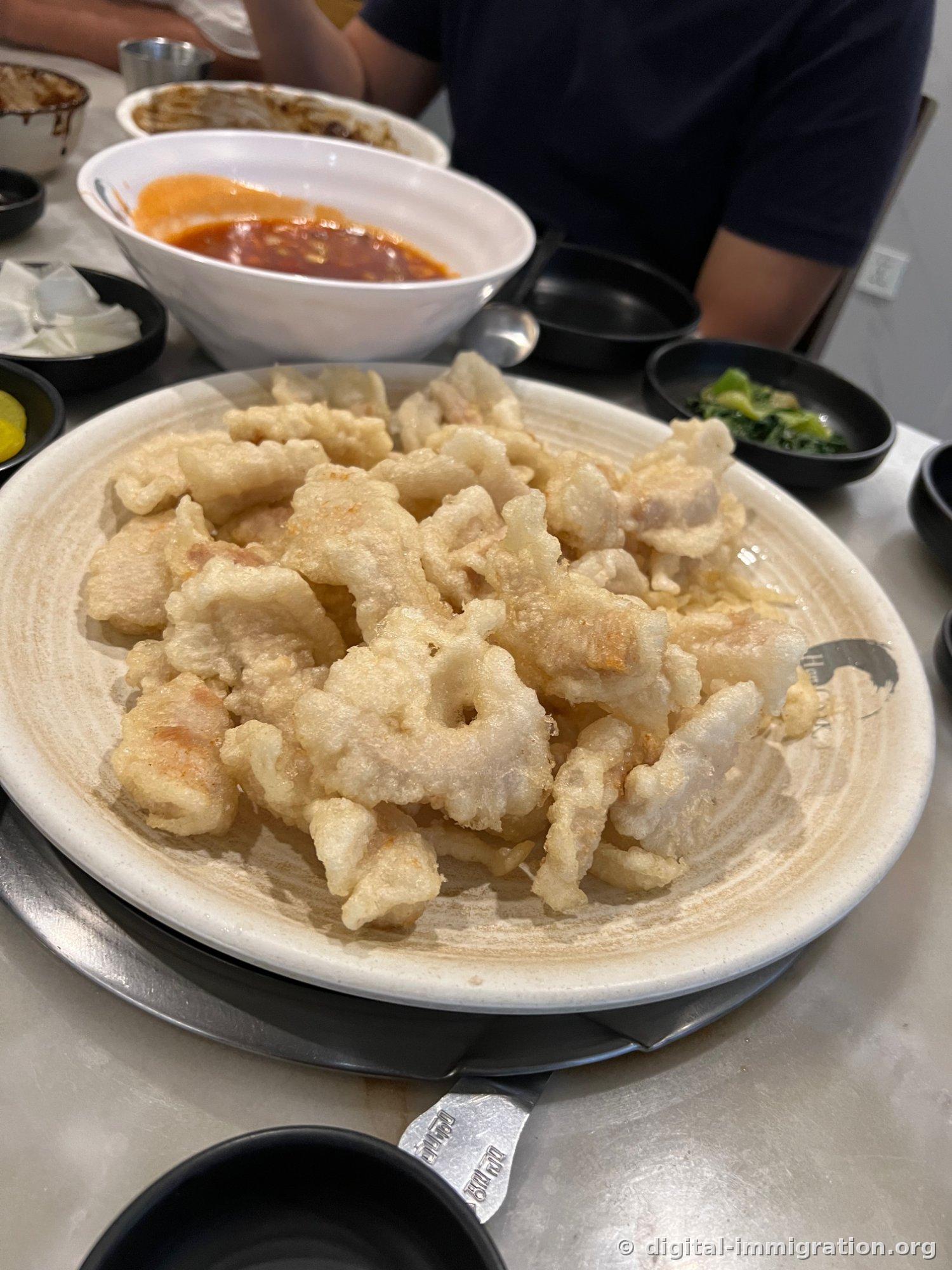 Tangsuyuk au riz gluant, à la pâte blanche et gonflée, servi en pile sur une assiette blanche portant le logo HANCOOK sur son pourtour. Derrière, un bol de jjamppong au bouillon rouge à moitié terminé, avec du danmuji et du pak-choï en accompagnement.