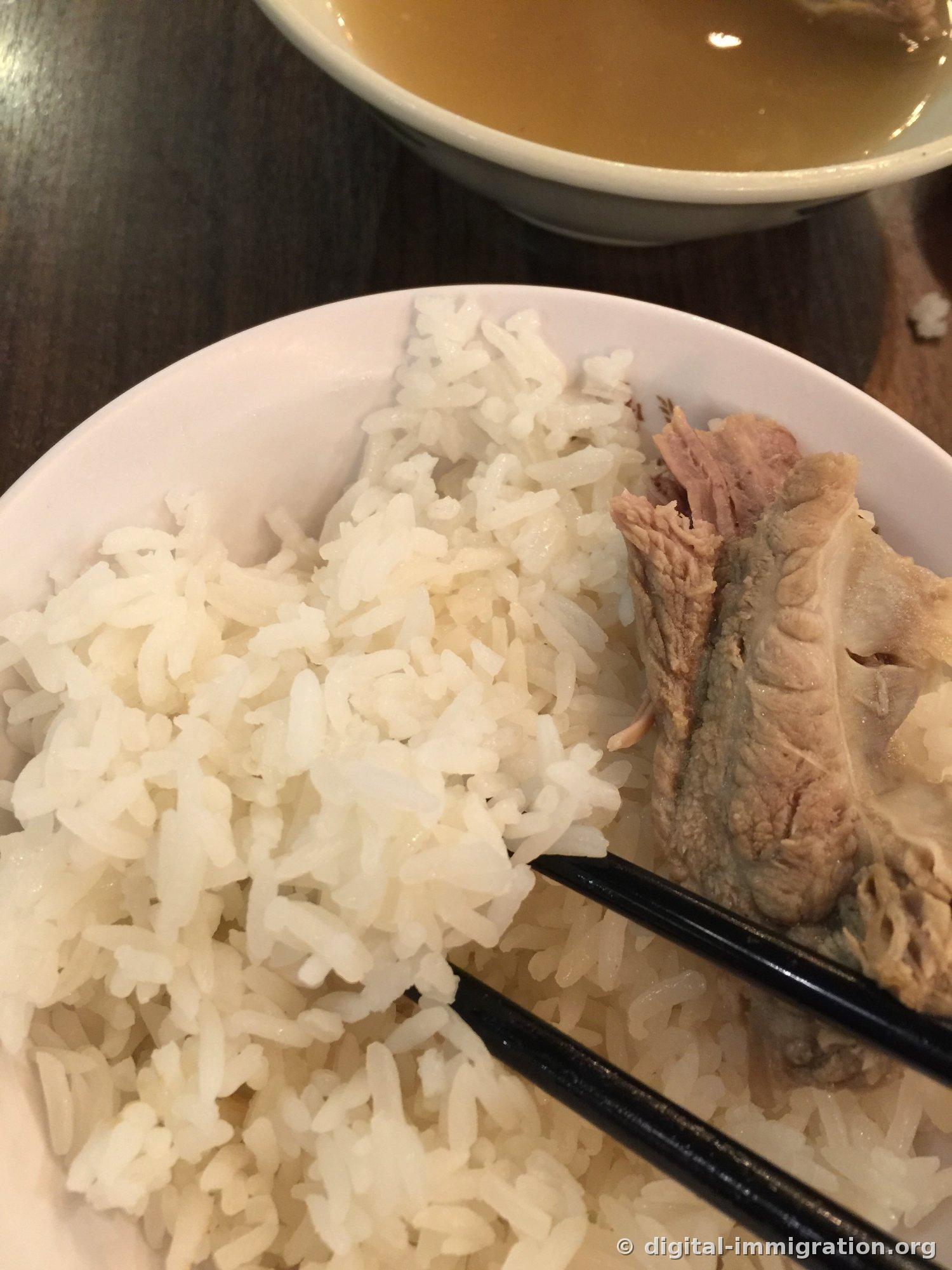 Un bol de riz blanc avec une côte de Bak Kut Teh posée dessus, pris avec des baguettes noires. Un bol de bouillon est visible derrière