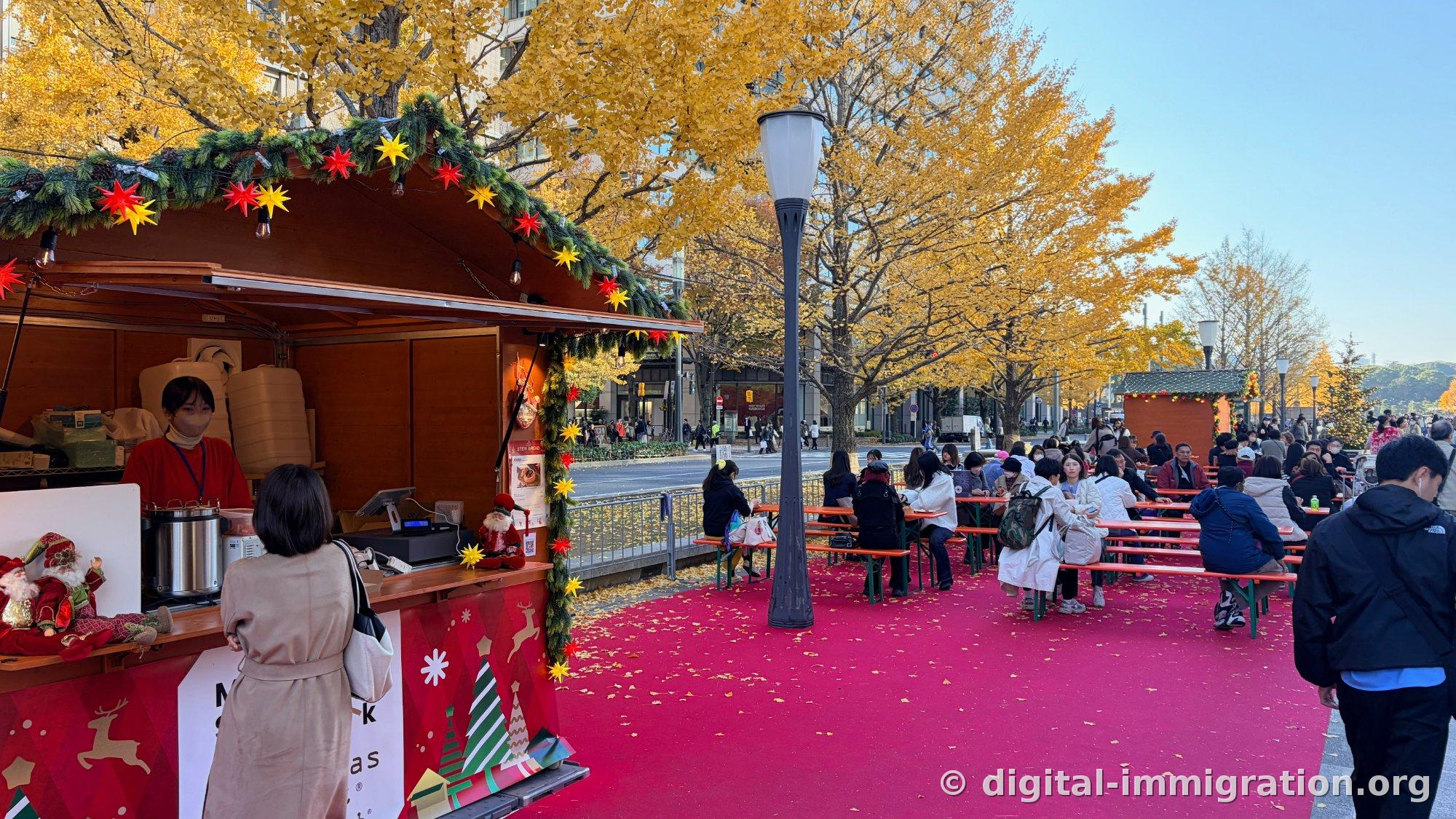 Le comptoir du chalet du marché de Noël avec sa grande cocotte-minute inox pour le vin chaud et les décoraciones étoilées, les tables de banquets pliables sur le sol rouge en arrière-plan et les ginkgos jaunes au-dessus