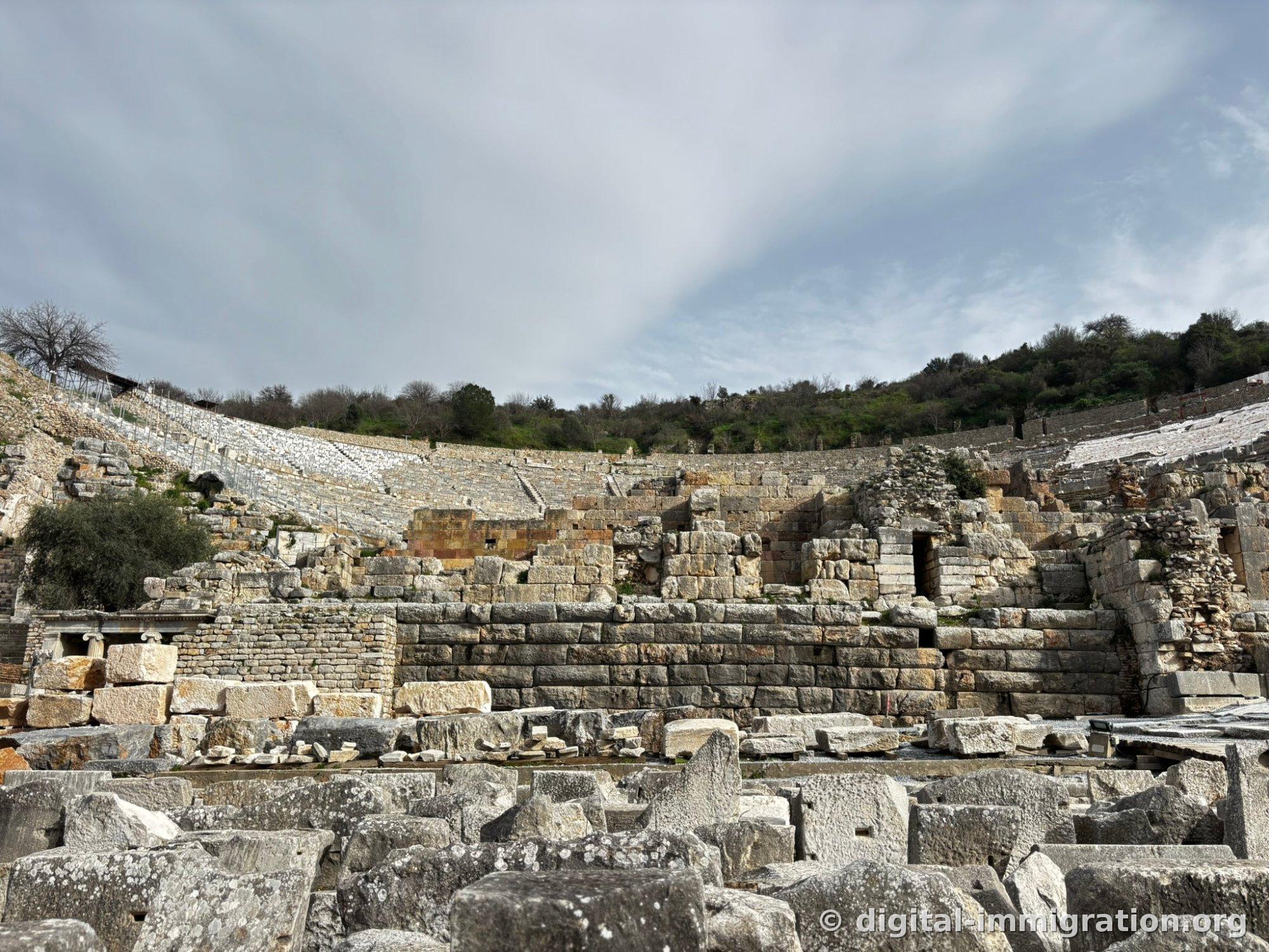 Ephesus Grand Theater carved into a hillside with its massive semicircular seating and collapsed stage building foundation in front, stone pieces scattered on the floor