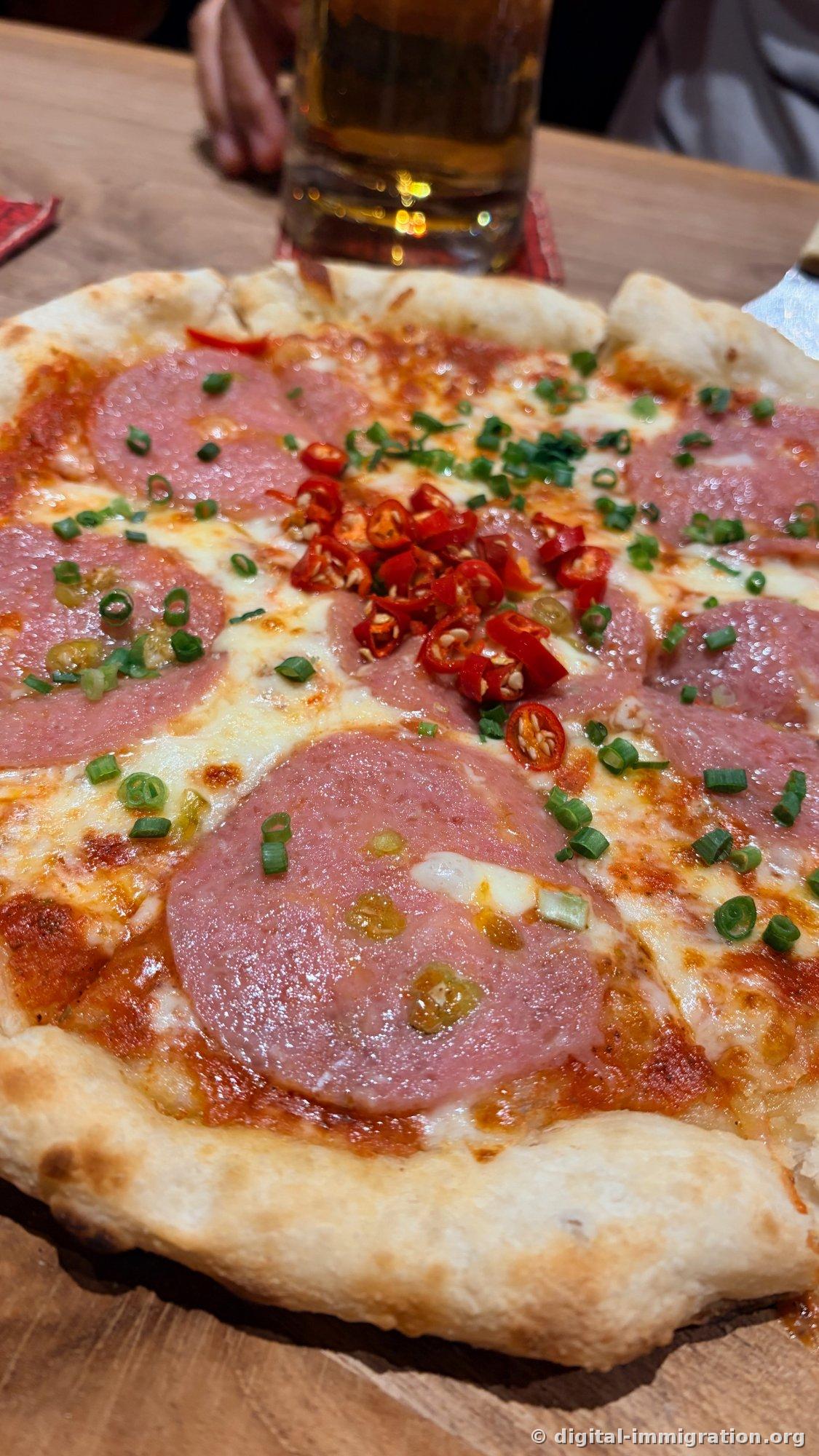 Une pizza fine avec du salami et de la mozzarella, garnie généreusement de tranches de piments rouges frais et d'oignons verts, un verre de bière derrière
