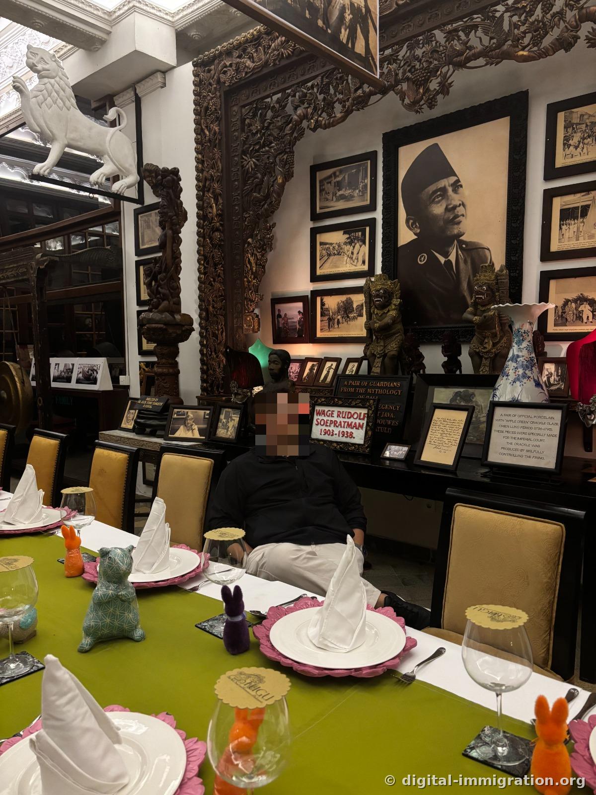 Dining table with green tablecloth and pink plate setting, behind it a wall with Sukarno portrait and Balinese guardian statues and old black and white photos