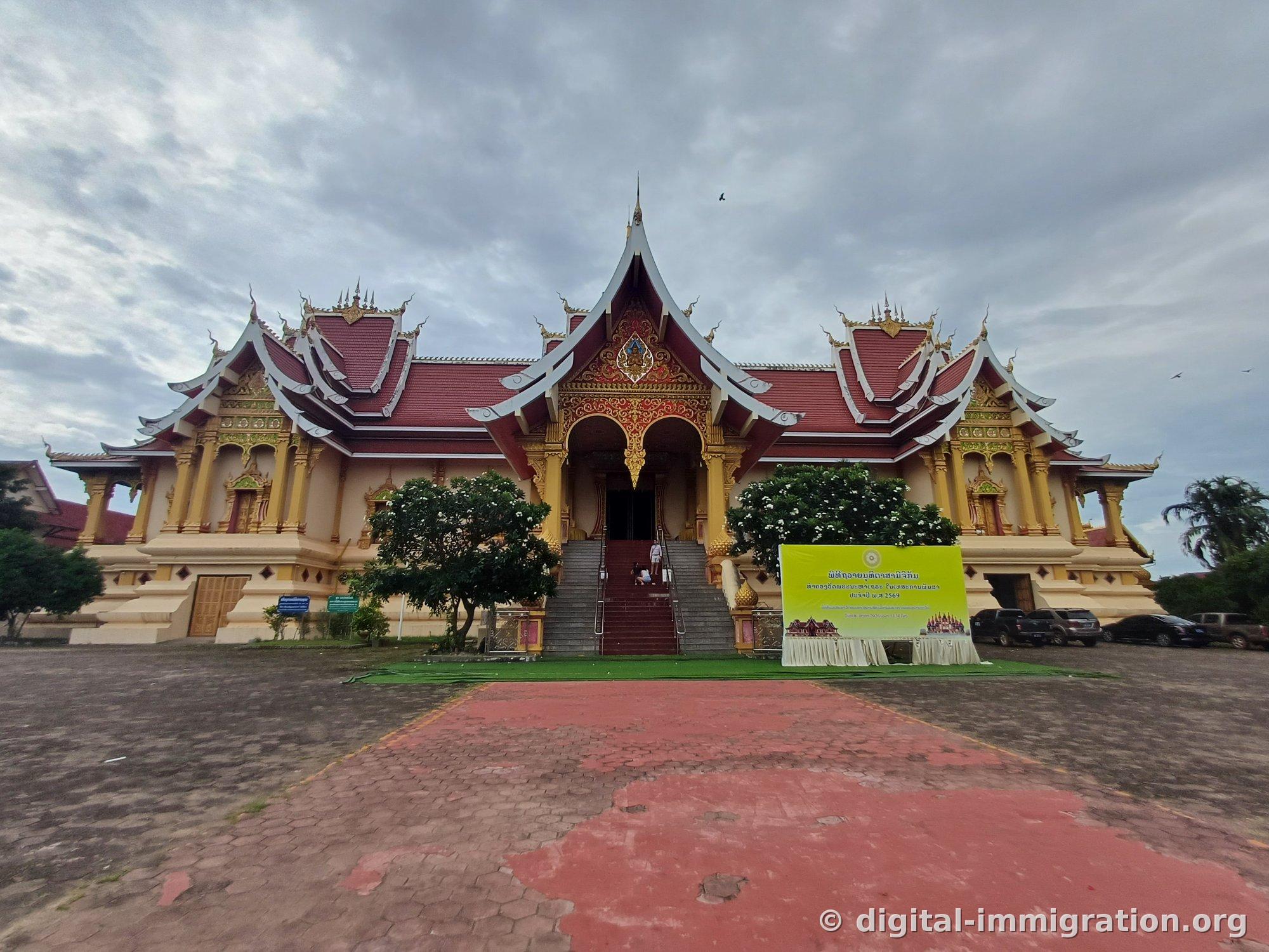 Laotian temple building with red tiled roof and gilded gable, yellow event banner, and artificial grass courtyard
