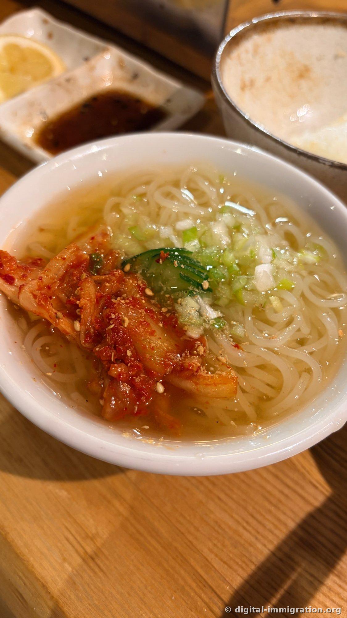 Cold noodles in a white bowl. Chopped green onion, a cucumber slice, and red kimchi are on top of the clear broth, with a lemon plate and beer glass nearby