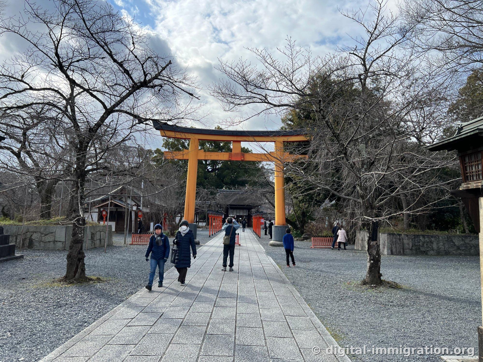 A large orange torii gate standing among bare cherry trees in winter, with a few worshippers walking the stone path through Hirano Shrine grounds