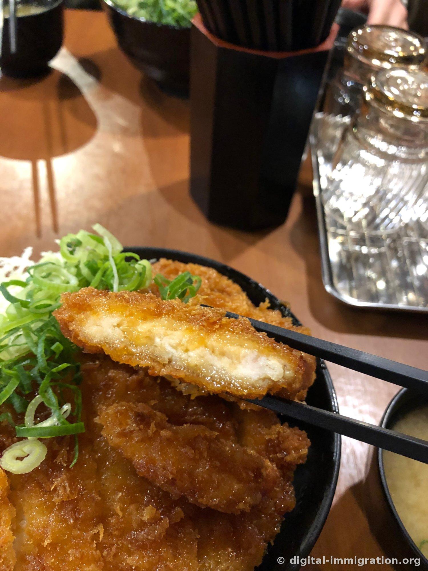 Section du katsu tenue avec des baguettes, la viande à l'intérieur de la panure apparaît blanche avec une bouteille d'eau et un porte-baguettes en arrière-plan