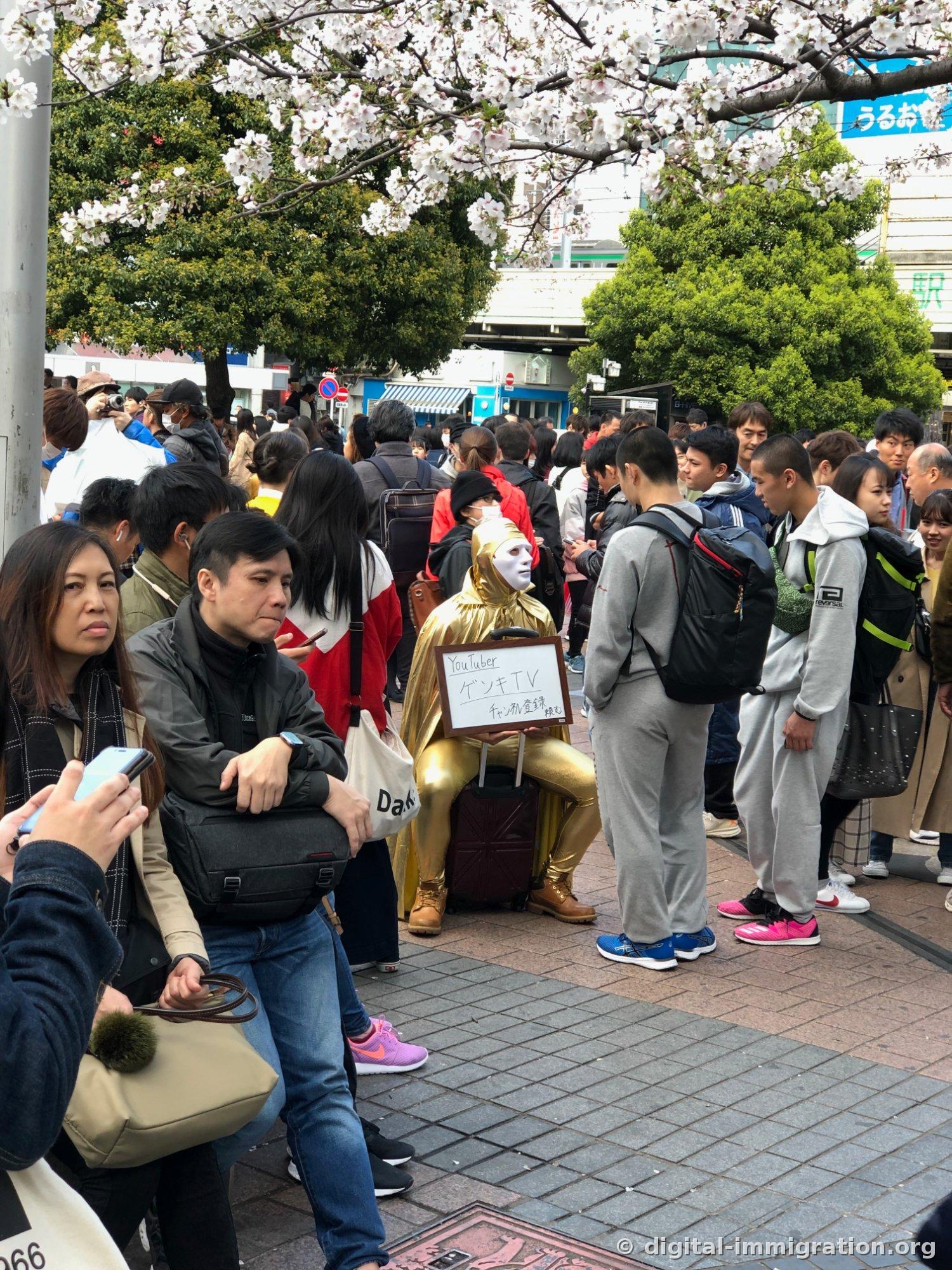 満開の桜の下、金色の全身タイツと白いお面姿で『YouTuber ゲンキTV』という札を持った人がキャリーバッグに座っている