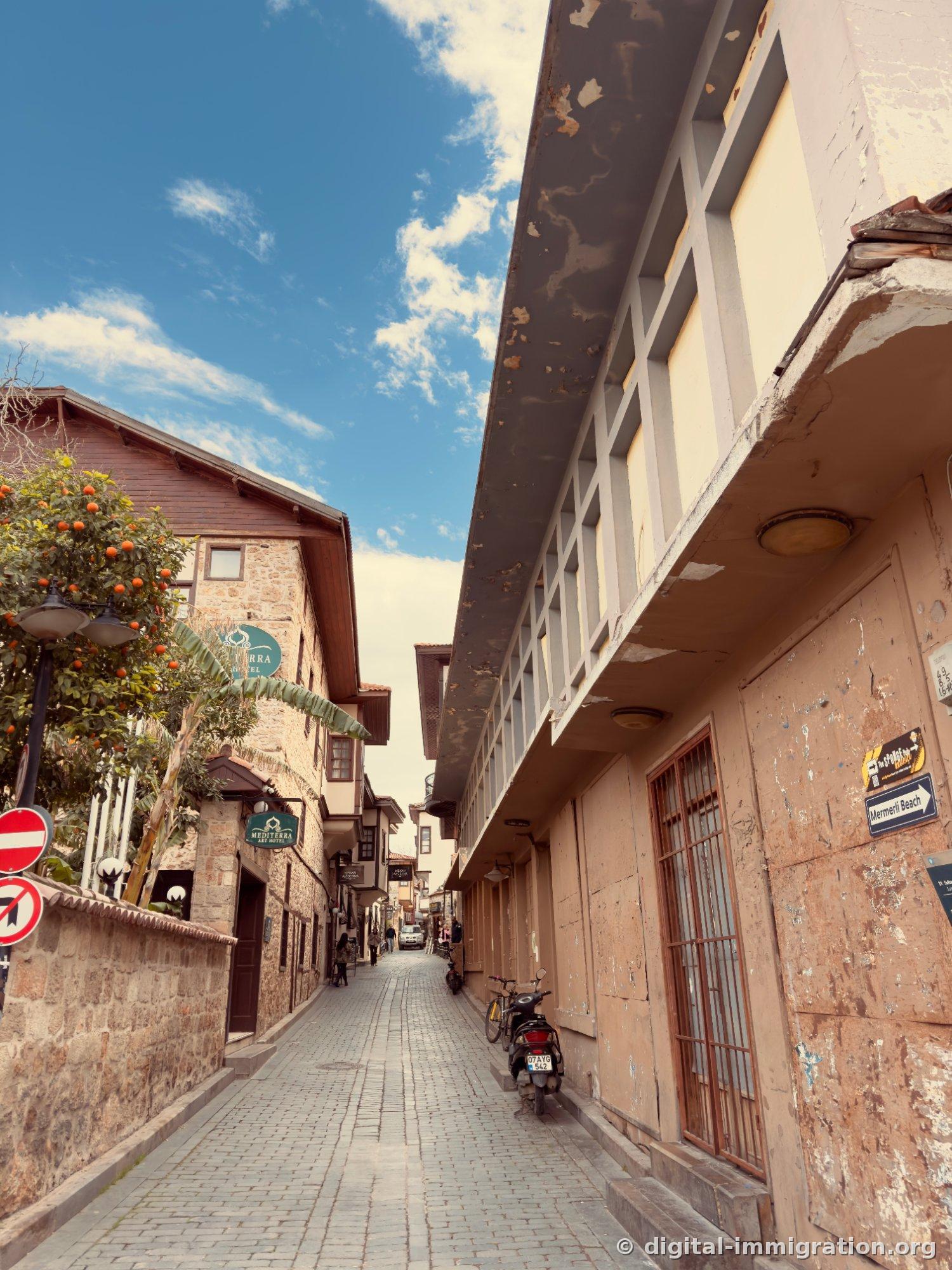 Ruelle étroite en pierre de Kaleiçi. À gauche, un oranger et l'enseigne du Mediterra Art Hotel, à droite, un panneau indiquant la direction de Mermerli Beach et un scooter garé contre le mur