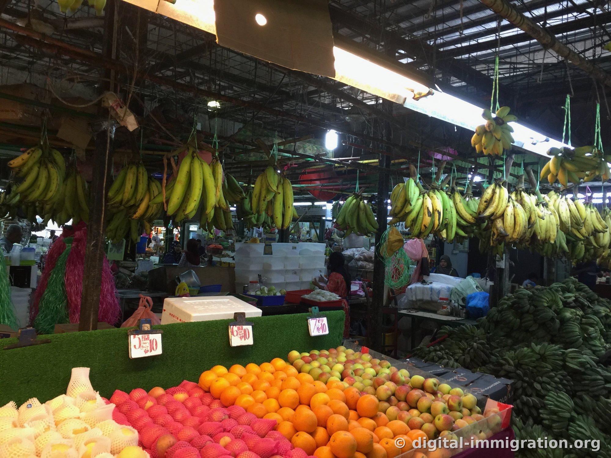 Banana bunches hanging in rows below steel ceiling and fluorescent lights, with pears, apples, and oranges in mesh bags displayed on stalls below