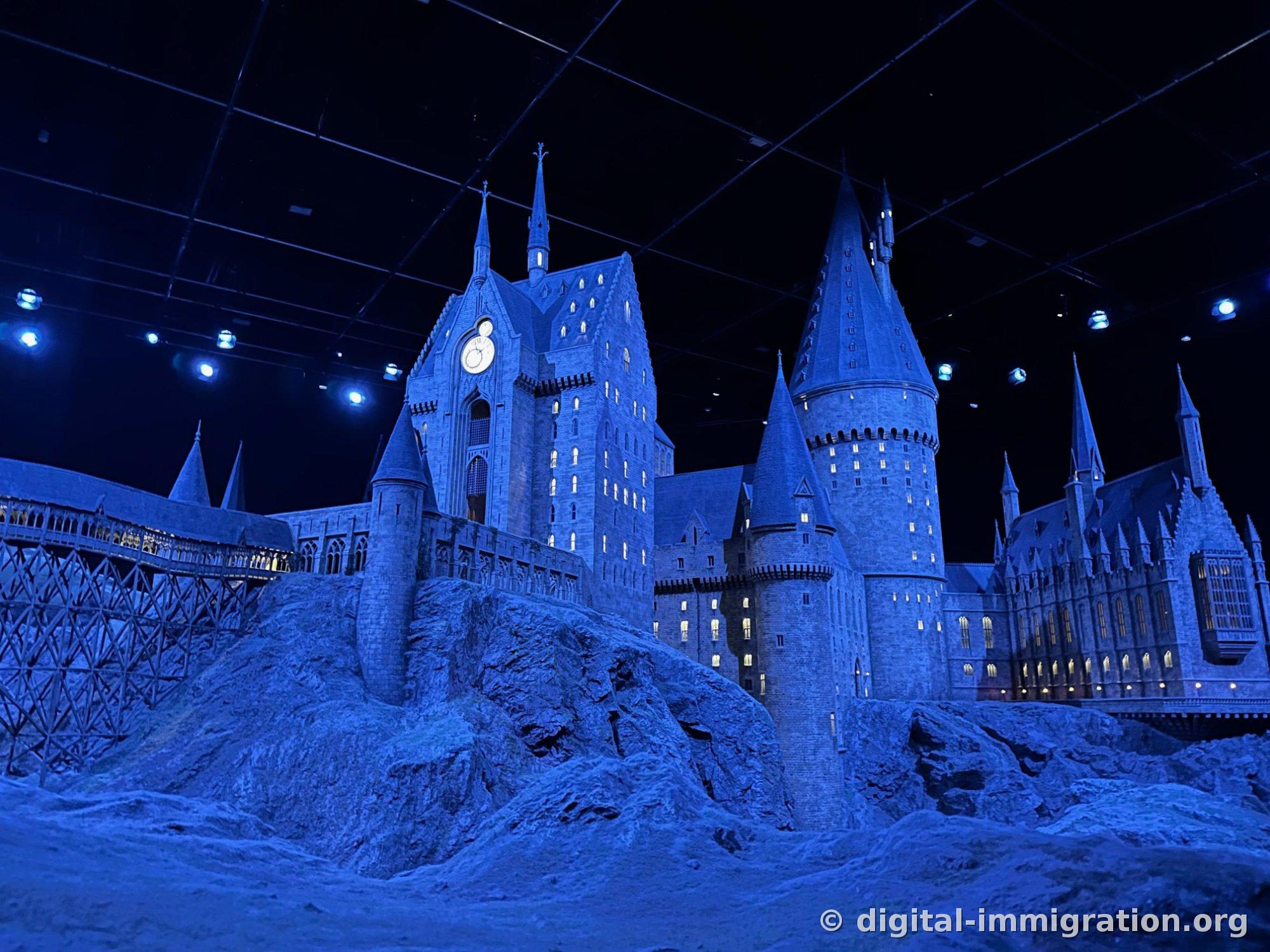 The same Hogwarts castle model under blue night lighting, with a wooden bridge visible on the left and the clock tower silhouetted