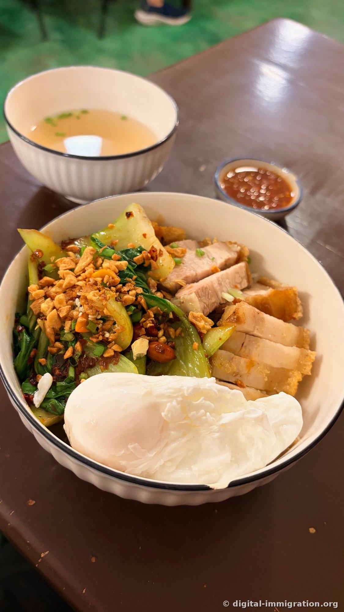 白い碗に炒めた青菜と挽いたピーナッツ、クリスピーポークのスライス、大きな温泉卵を乗せた丼。奥に透明なスープと唐辛子ダレの磁器の鉢が置かれている