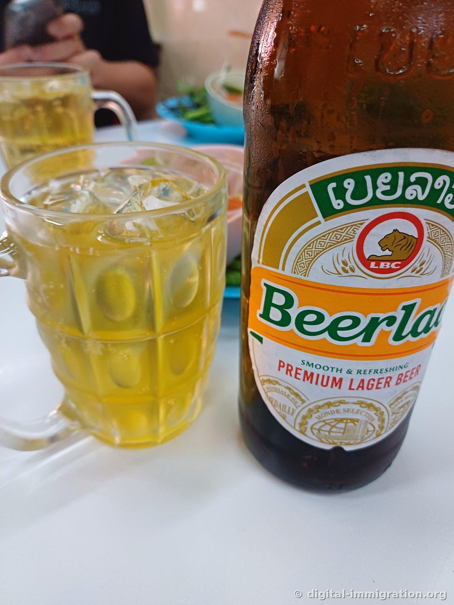 餐桌上水珠满布的Beerlao褐色啤酒瓶和装满冰块的有手柄玻璃啤酒杯，背后是蓝色盘子里的蔬菜