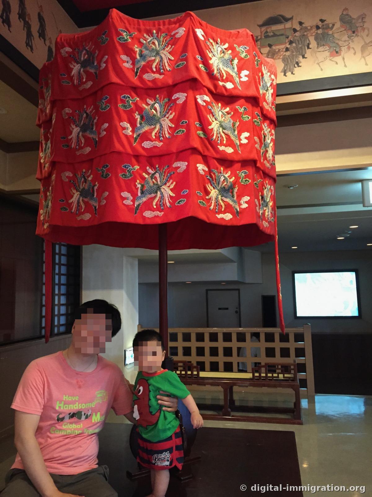 An adult and child sitting under the red royal canopy with phoenix and cloud embroidery, king's procession paintings on the wall behind