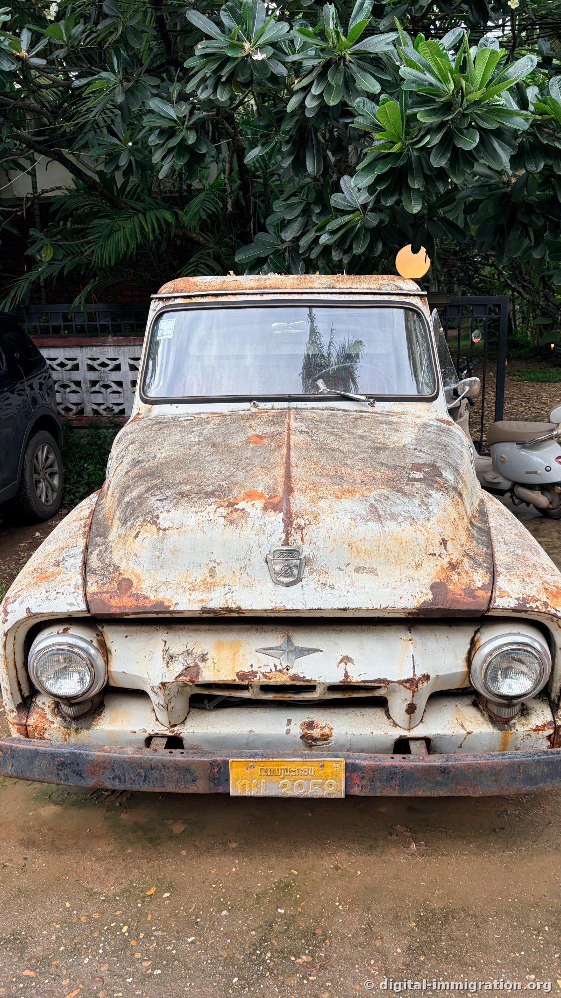 Un vieux pickup Ford blanc garé de face sous un fleur de plumeria, la peinture écaillée avec la rouille qui s'étend, une plaque d'immatriculation jaune du Laos à l'avant
