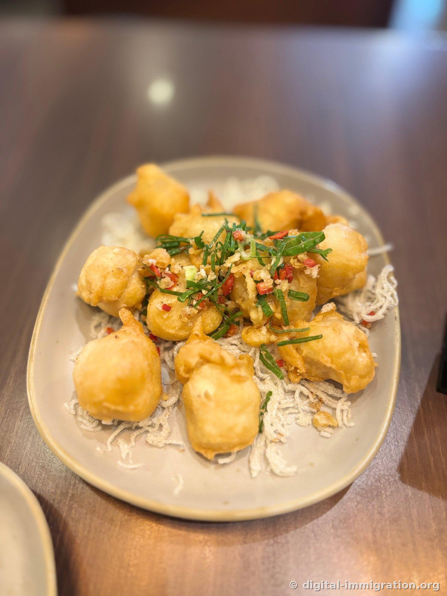 Chunks of fried soft tofu placed on a bed of fried noodles, topped with minced garlic, red chili, and sliced scallions