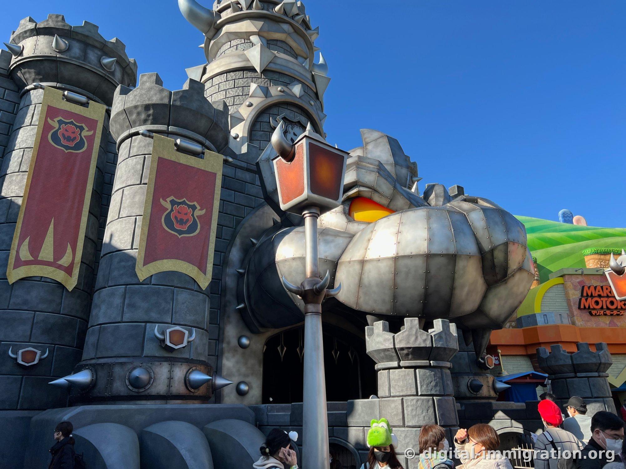The outer wall of Bowser's Castle made of gray stone, a red banner with Bowser's crest, and a silver metal Bowser sculpture protruding above the castle wall