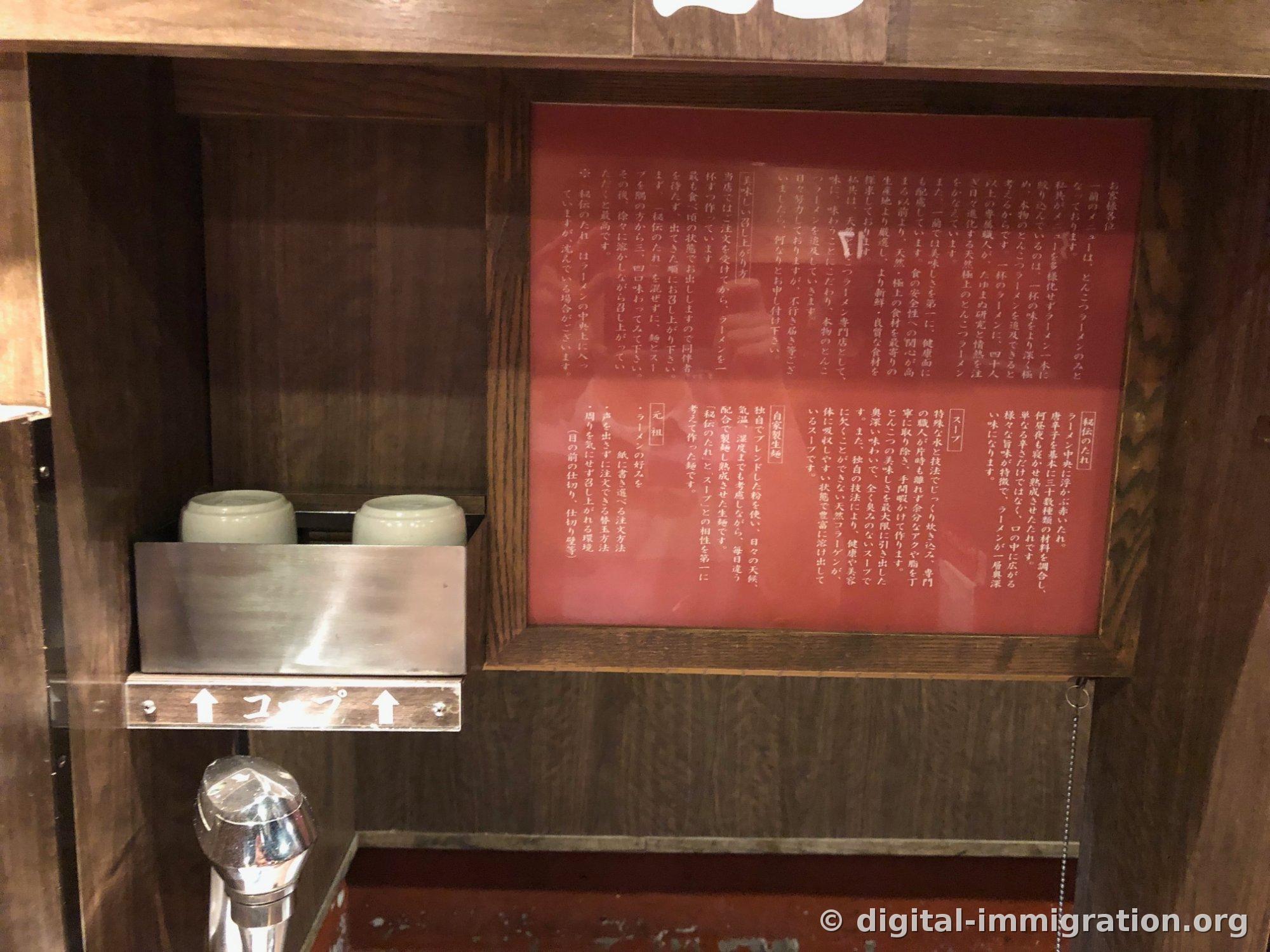 Sur le mur du comptoir, deux tasses et un robinet à appuyer, avec à côté un cadre rouge contenant les explications sur la sauce spéciale et le bouillon