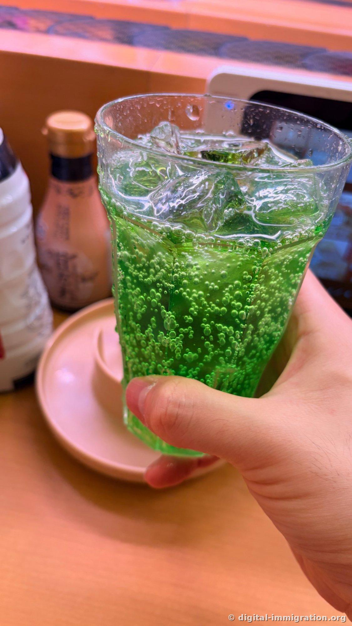 Un verre de soda vert fluorescent rempli de glaçons et de bulles tenu à la main, avec une bouteille de sauce soja et une table en bois visibles à l'arrière-plan