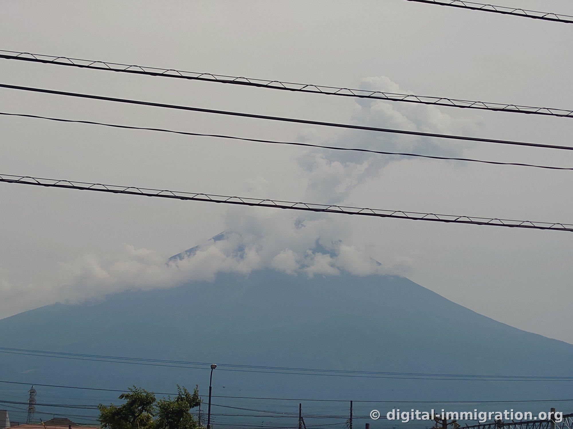 電線の向こうに富士山の頂上から白い雲が煙のように垂直に立ち昇っている様子を望遠で撮った写真