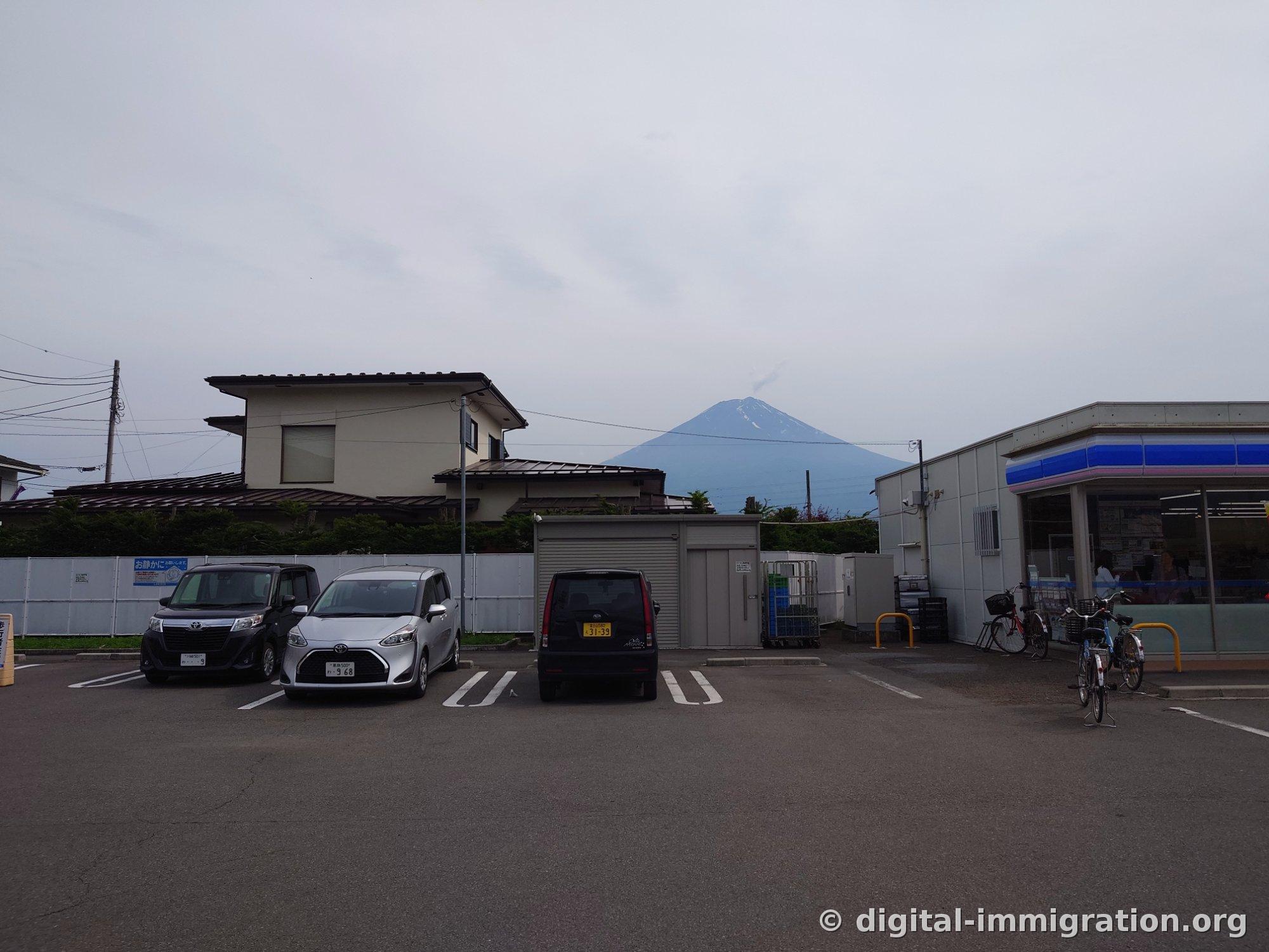 A quiet convenience store parking lot with small cars and bicycles parked, and Mount Fuji with lingering patches of snow visible beyond the rooftops on a cloudy day