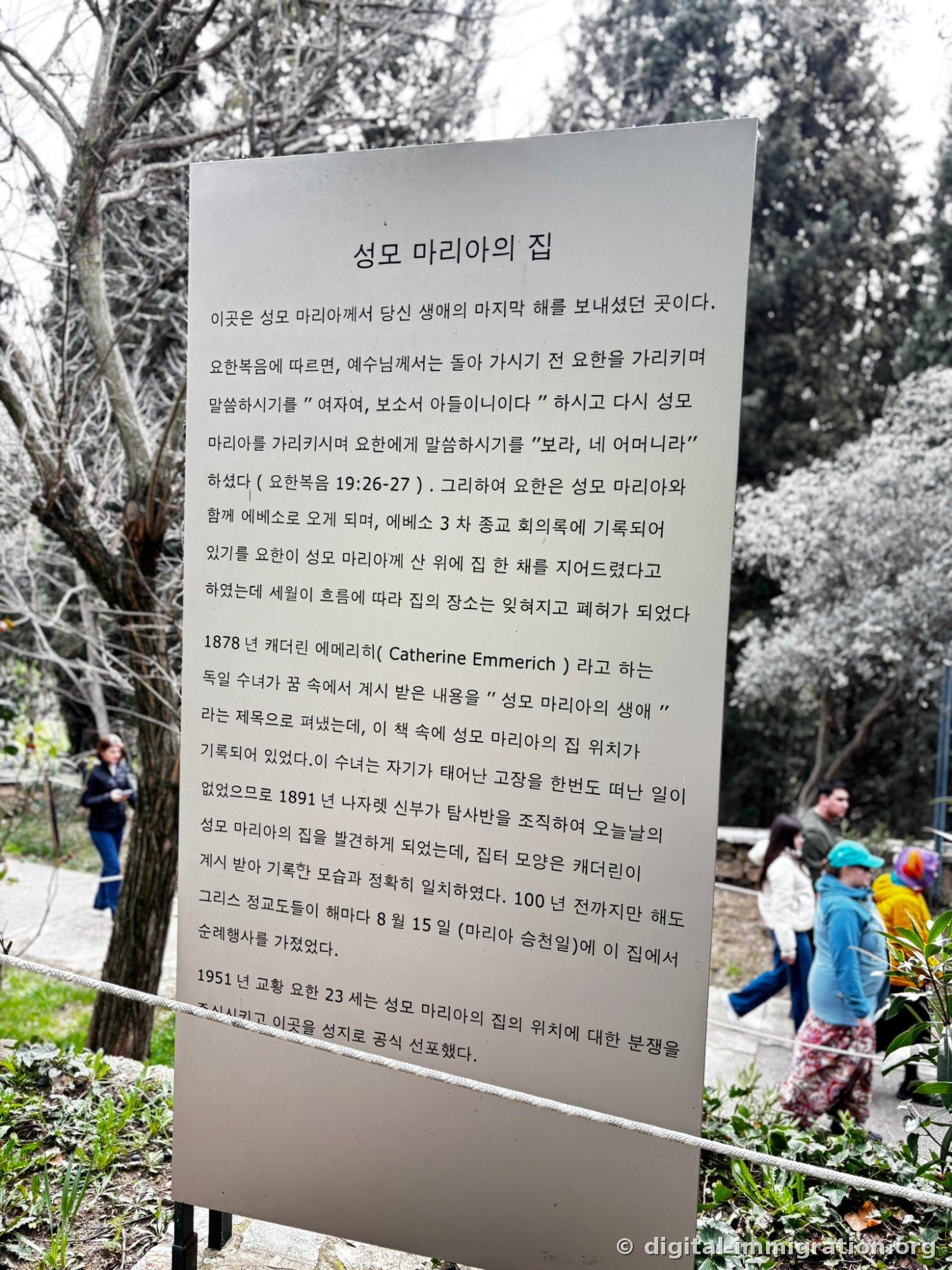 A Korean information plaque titled 'House of the Virgin Mary' stands among trees with visitors walking past in the background