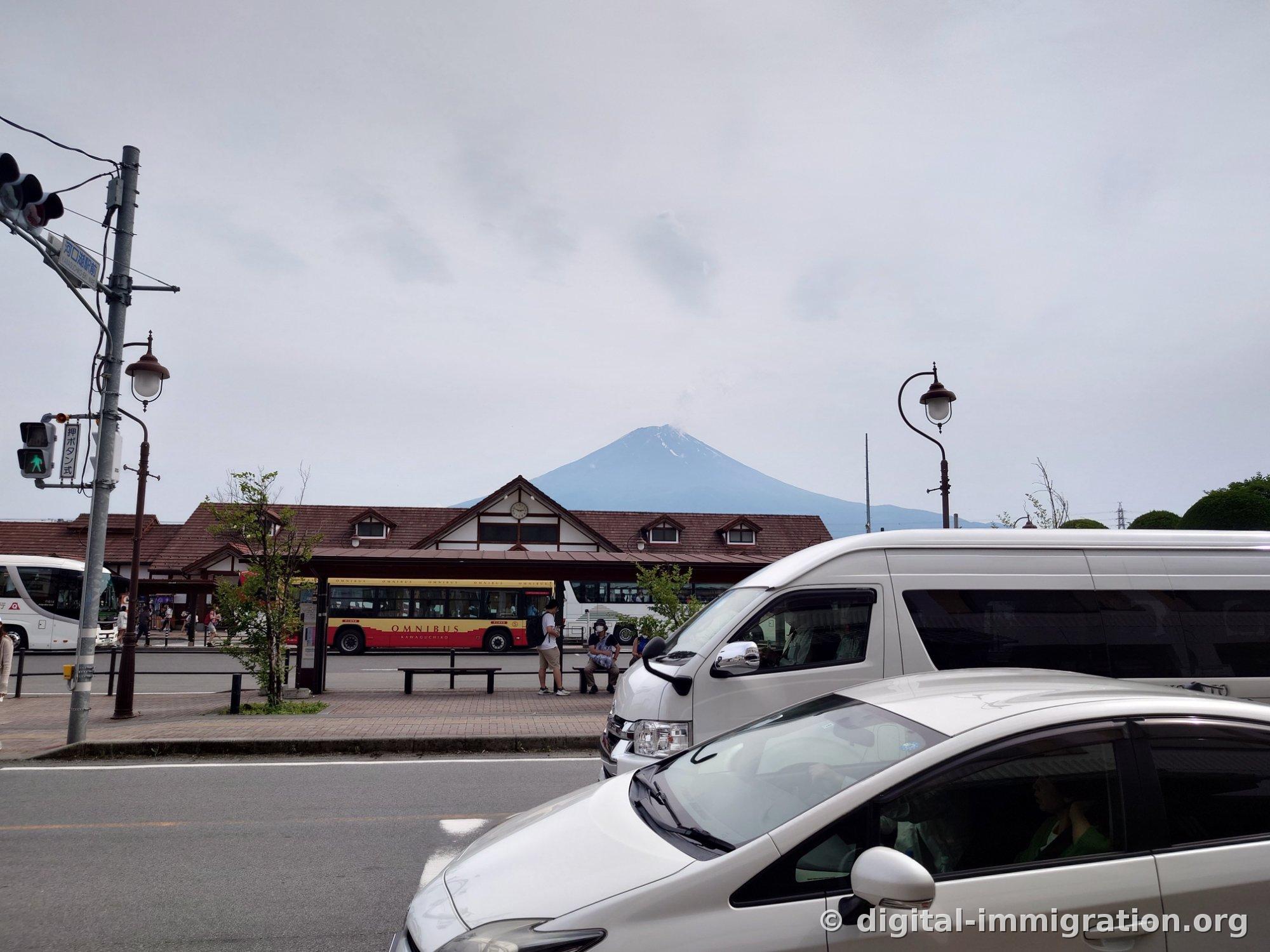Behind the triangular-roofed Kawaguchiko Station building, Mount Fuji's peak rises above, with a red and yellow Kawaguchiko Omnibus tour bus waiting at the stop