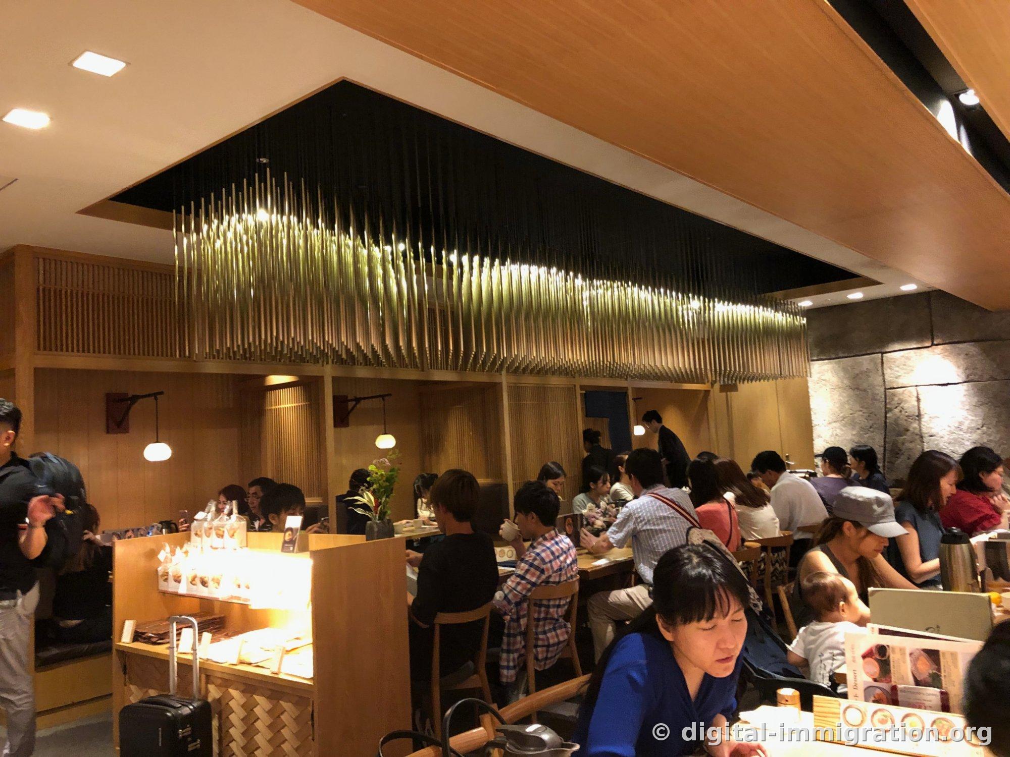 Interior of the Tsurutontan hall full of customers under bamboo-suspended ceiling lighting, with luggage standing on the left