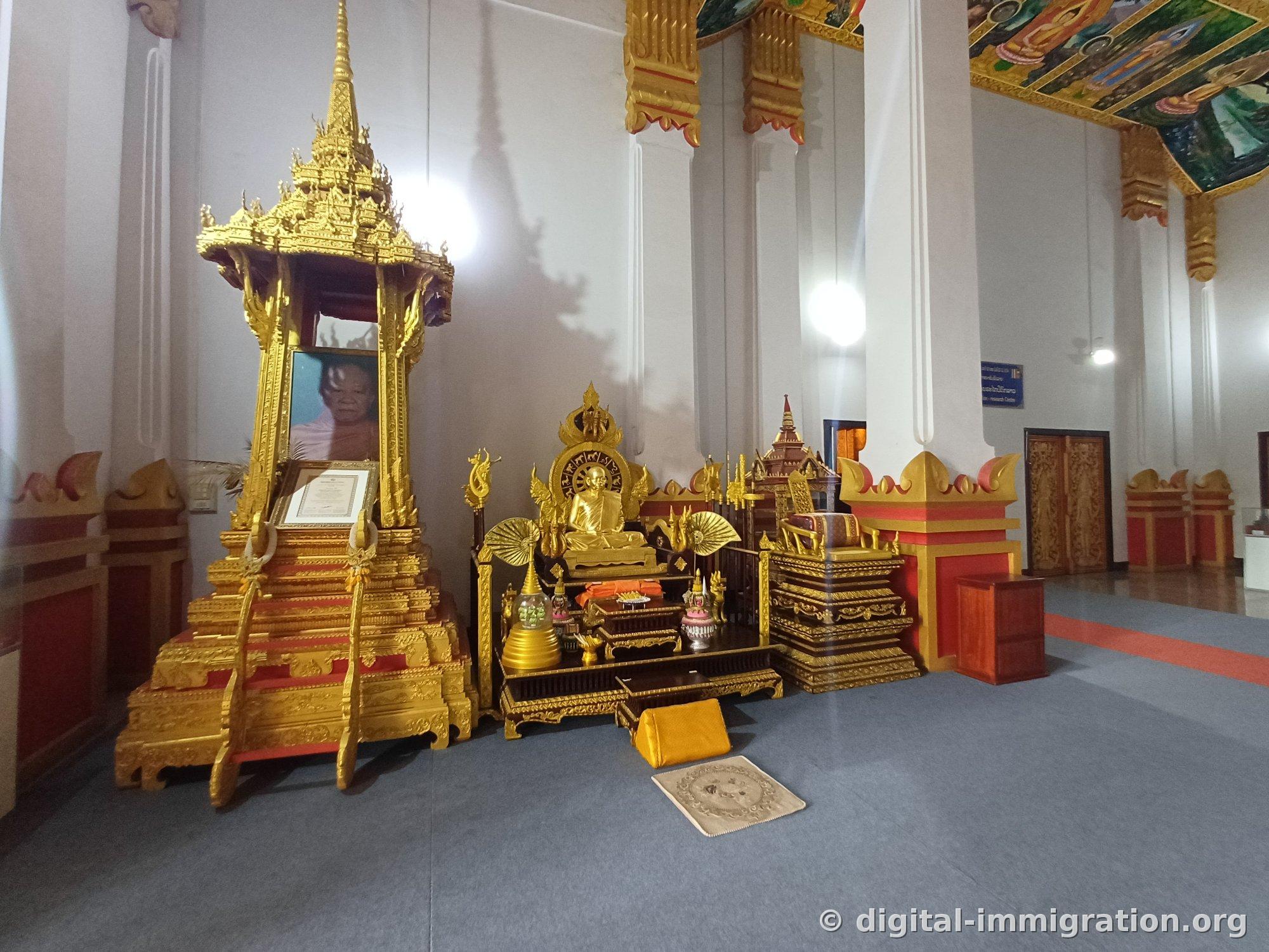Portrait and framed documents enshrined in a golden pavilion with golden statue, fan, and prayer mats beside it