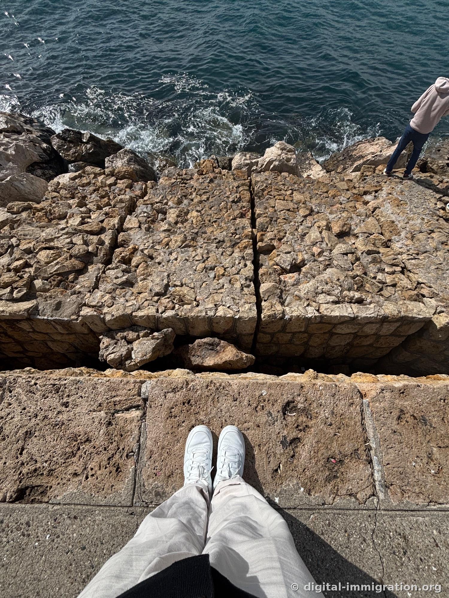First-person view of feet at the harbor stone retaining wall edge, deep blue seawater splashing through cracks, person standing on rocks at upper right