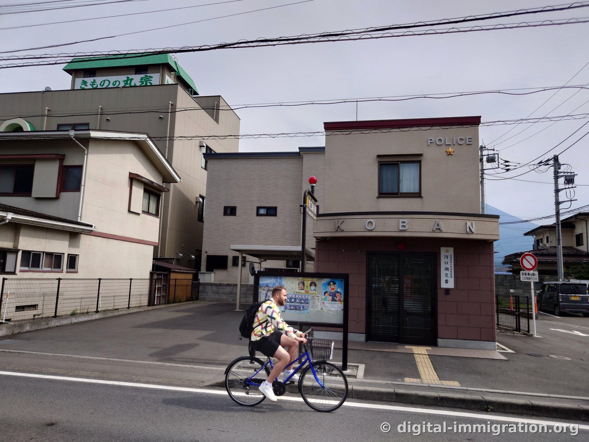 A traveler on a blue bicycle passes in front of the Kawaguchiko police box building with its KOBAN sign, with a kimono shop sign visible on the left