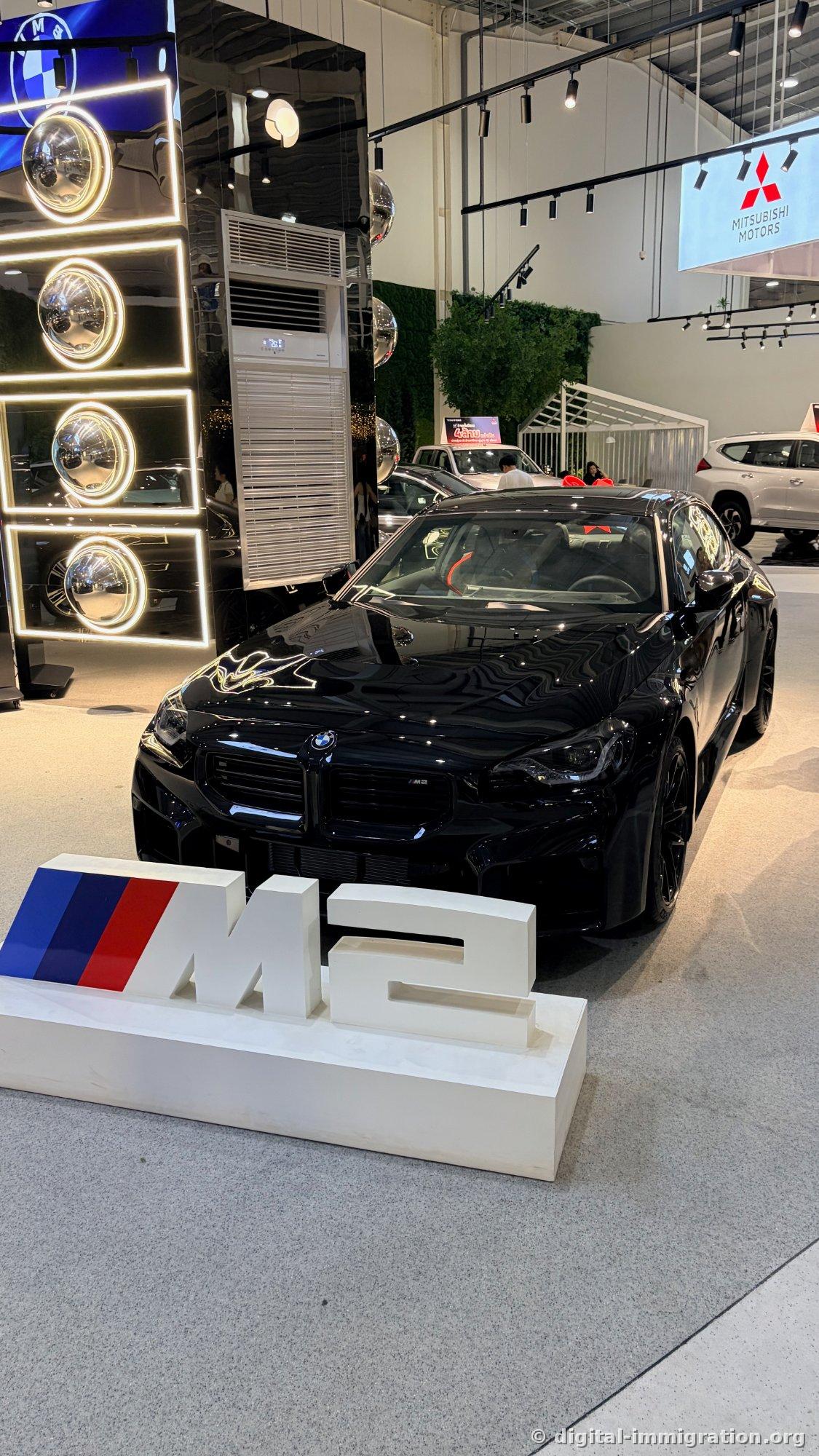M2 logo sculpture on white base with black BMW M2 coupe behind, MITSUBISHI MOTORS sign on upper right
