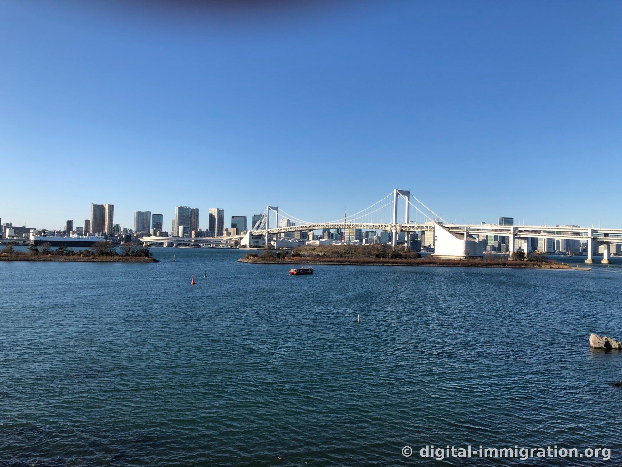 从台场甲板看东京湾全景，白色彩虹大桥和市中心高楼天际线，水面上有一艘红色屋形船
