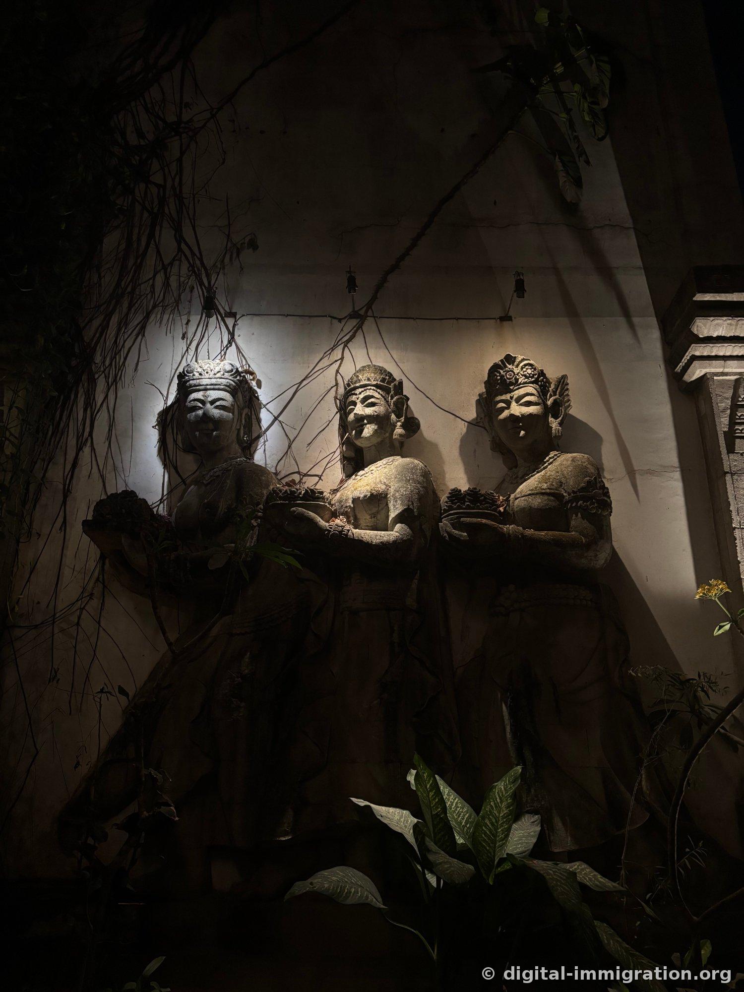 Three stone female statues in a row supporting offering vessels on a dark wall, dry vines hanging above covering their faces
