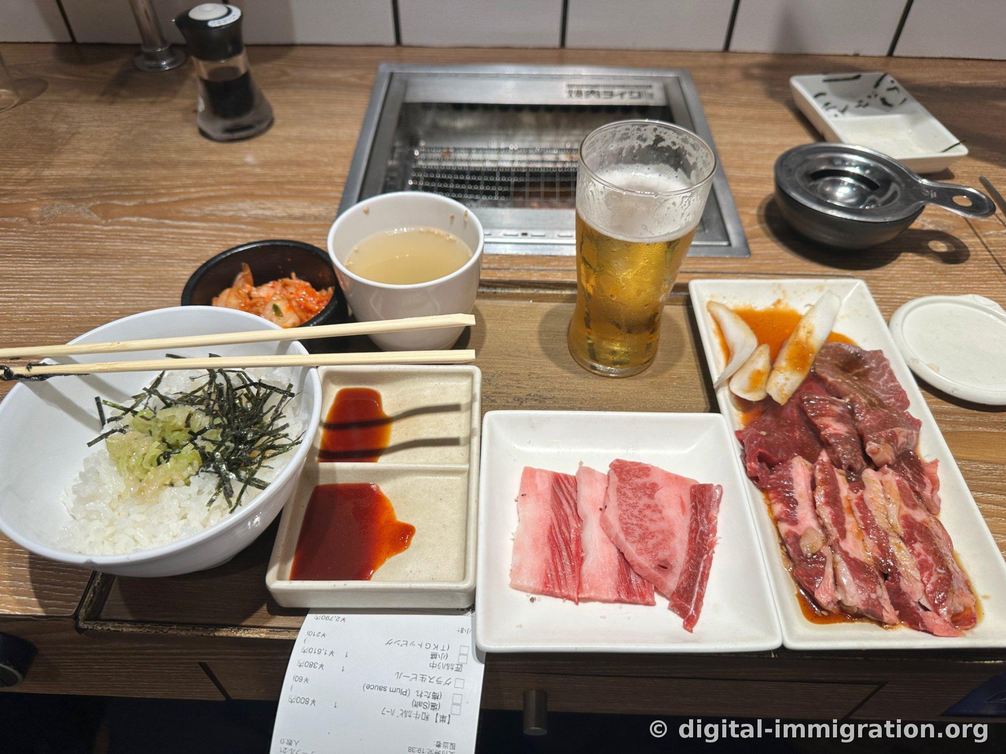 Un foyer individuel posé sur un comptoir en bois, avec du riz aux œufs et sauce soja, du kimchi, une soupe, une bière fraîche, une assiette de sauces, deux assiettes de viande crue, et le reçu