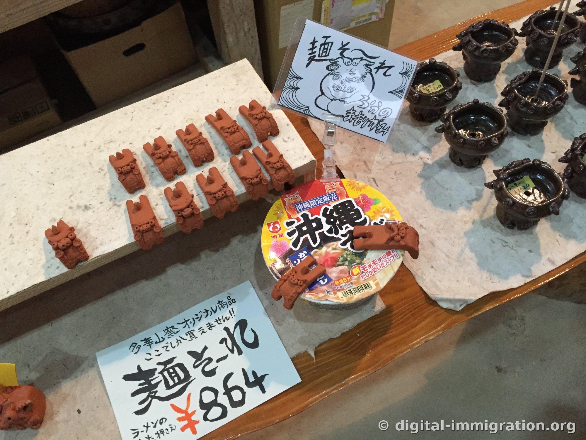 Sur l'étal de vente : des presse-couverles de ramen en forme de lion Shisa alignés avec un panneau manuscrit « 麺そ〜れ ¥864 », une exposition de démonstration avec un bol de ramen instantané