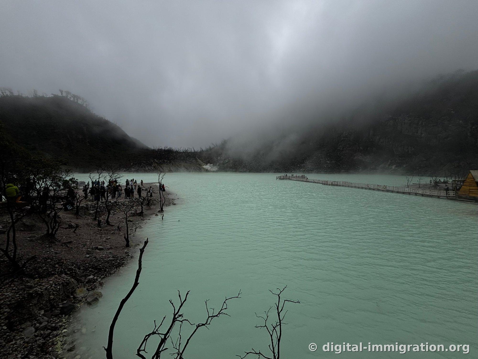 Inside the misty crater, people gathered on the gravel beach on the left side of the cyan lake, with a long wooden deck and yellow hut extending to the right