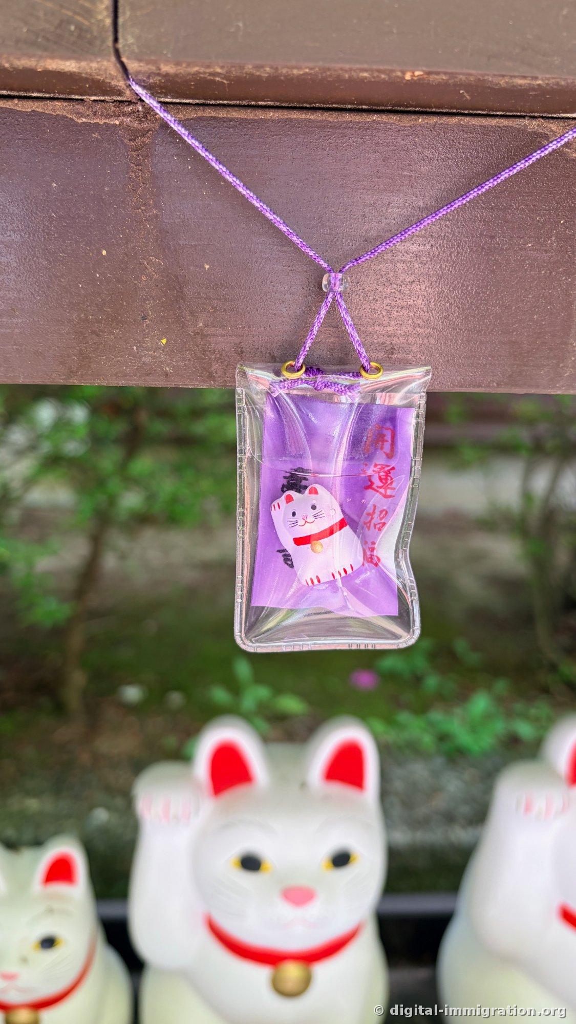 A purple omamori with a beckoning cat and 'Kaiunshofuku' printed on it, hanging from a wooden post with cats in the background