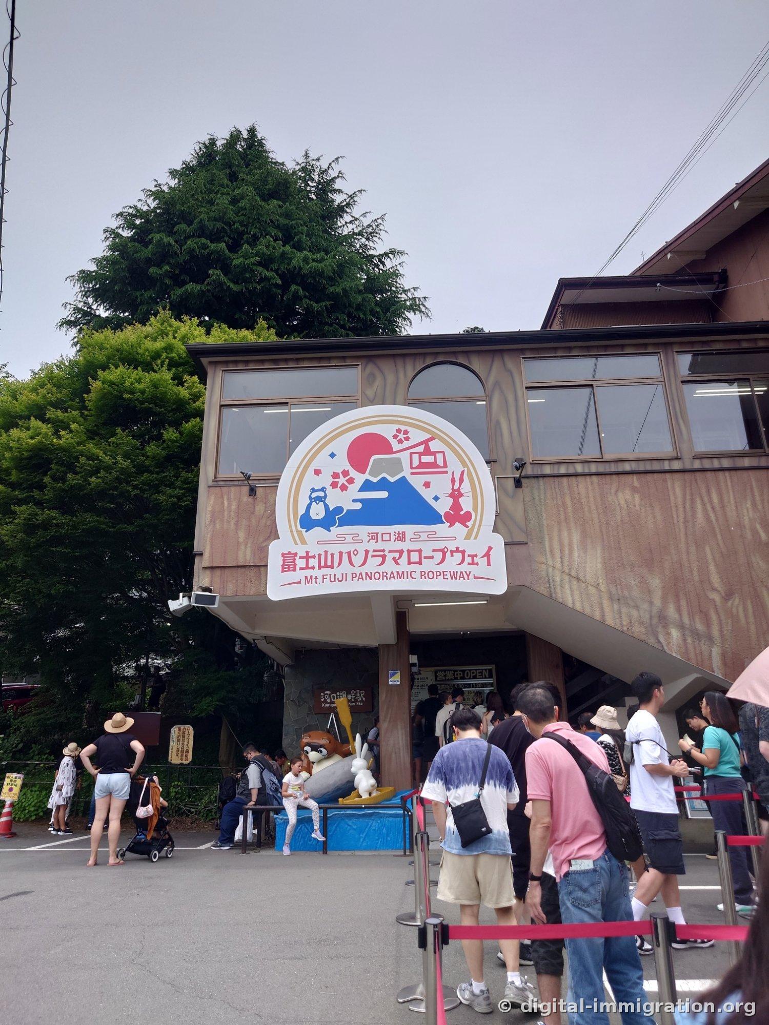 The circular '富士山パノラマロープウェイ' sign hanging on the Kawaguchiko Sanbasaki Station building and people in line below it