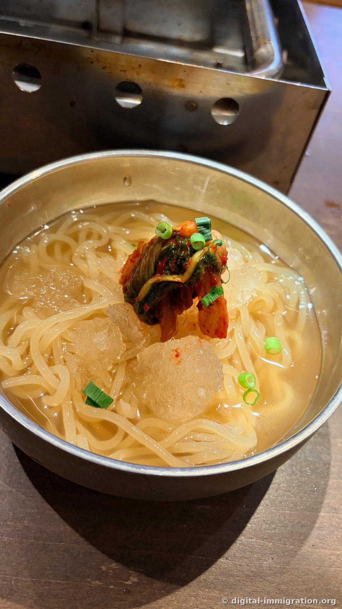 Cold noodles in a stainless-steel bowl; large ice chunks float on the broth with napa cabbage kimchi and chopped scallions in the center